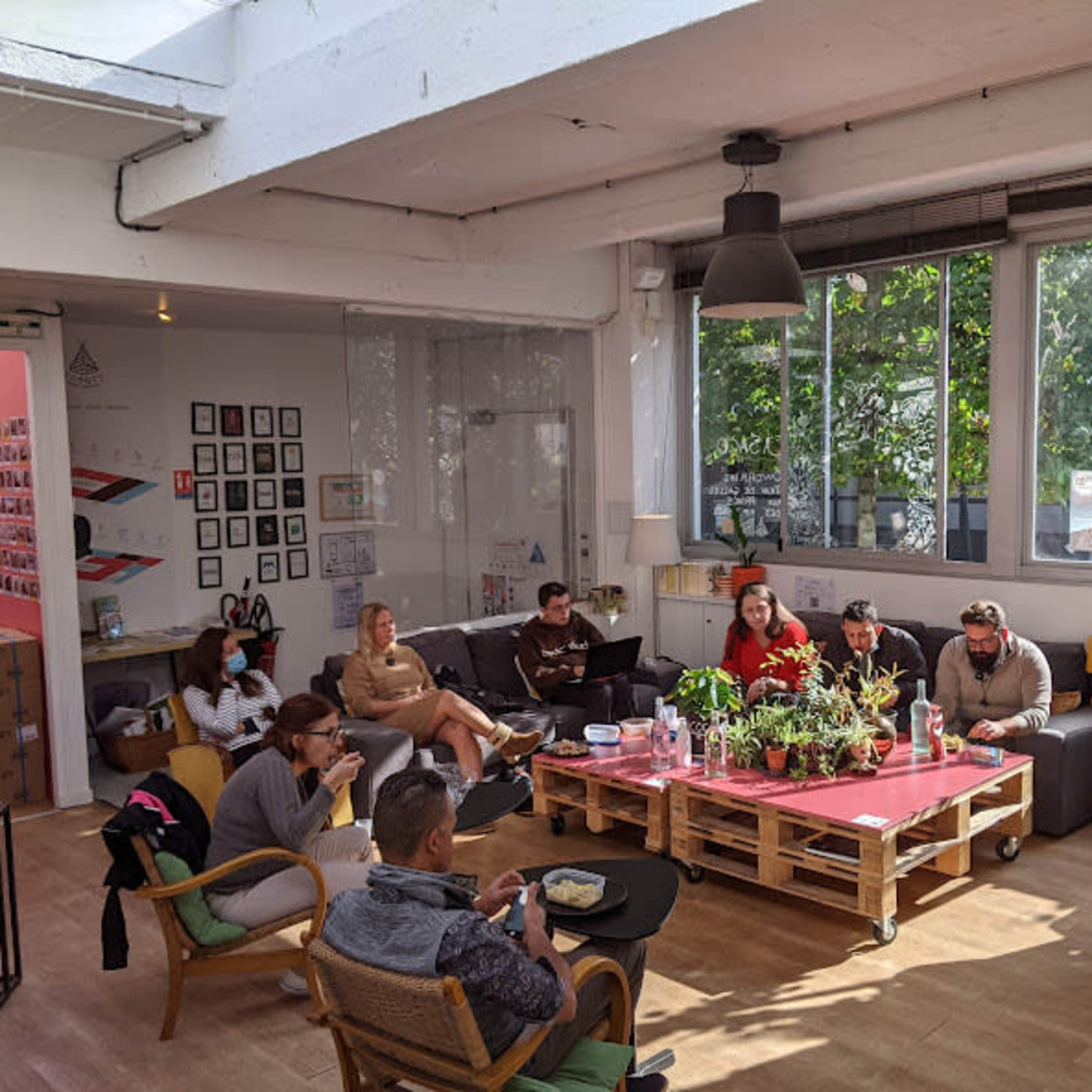 A group of people sits in a bright, open room with large windows, surrounded by plants and casual seating.