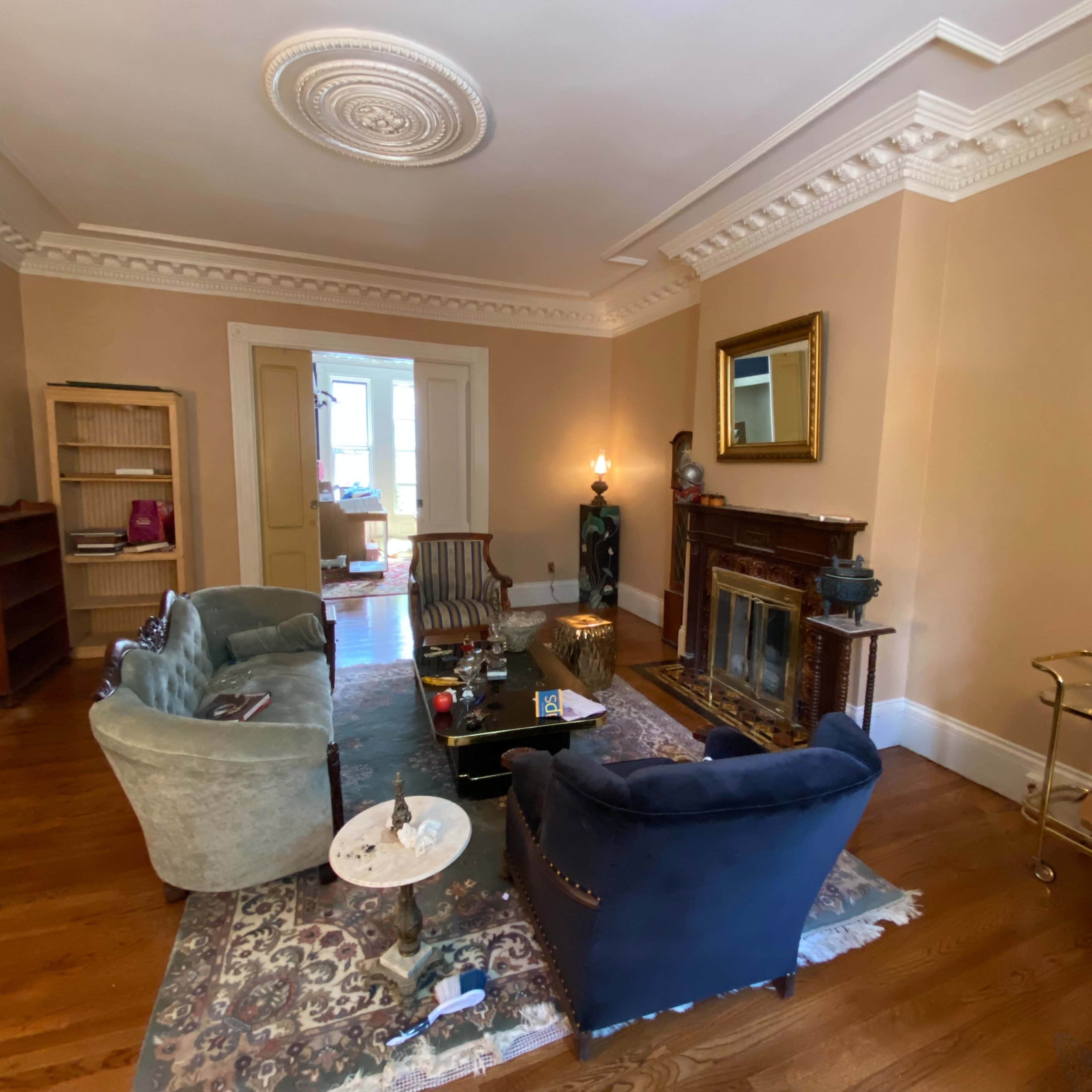 A living room features two upholstered sofas, a coffee table, a fireplace with a mirror above it, a small bookshelf, and a bar cart in a historic setting.