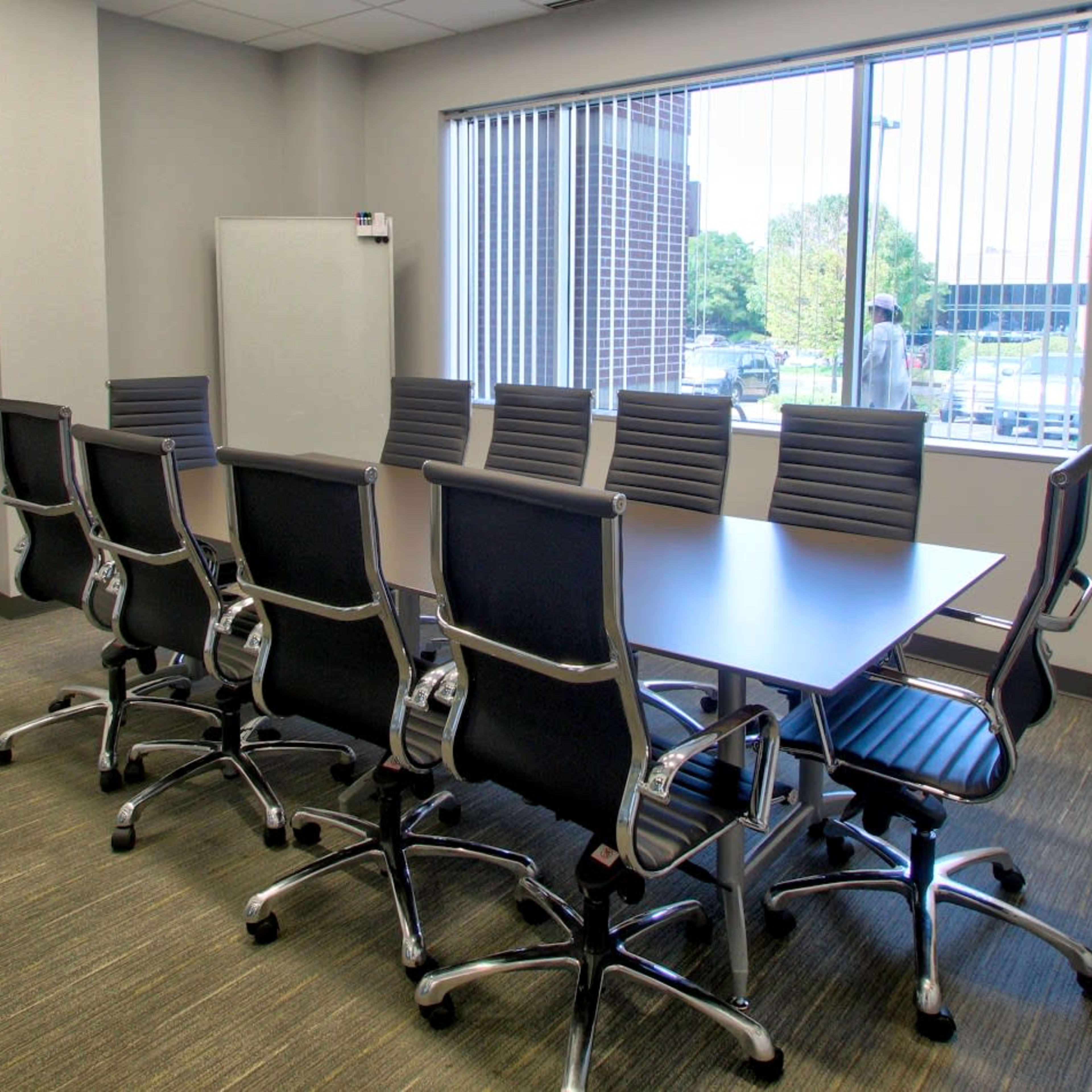 A conference room features a long table surrounded by eight ergonomic chairs and a window overlooking an outdoor view.