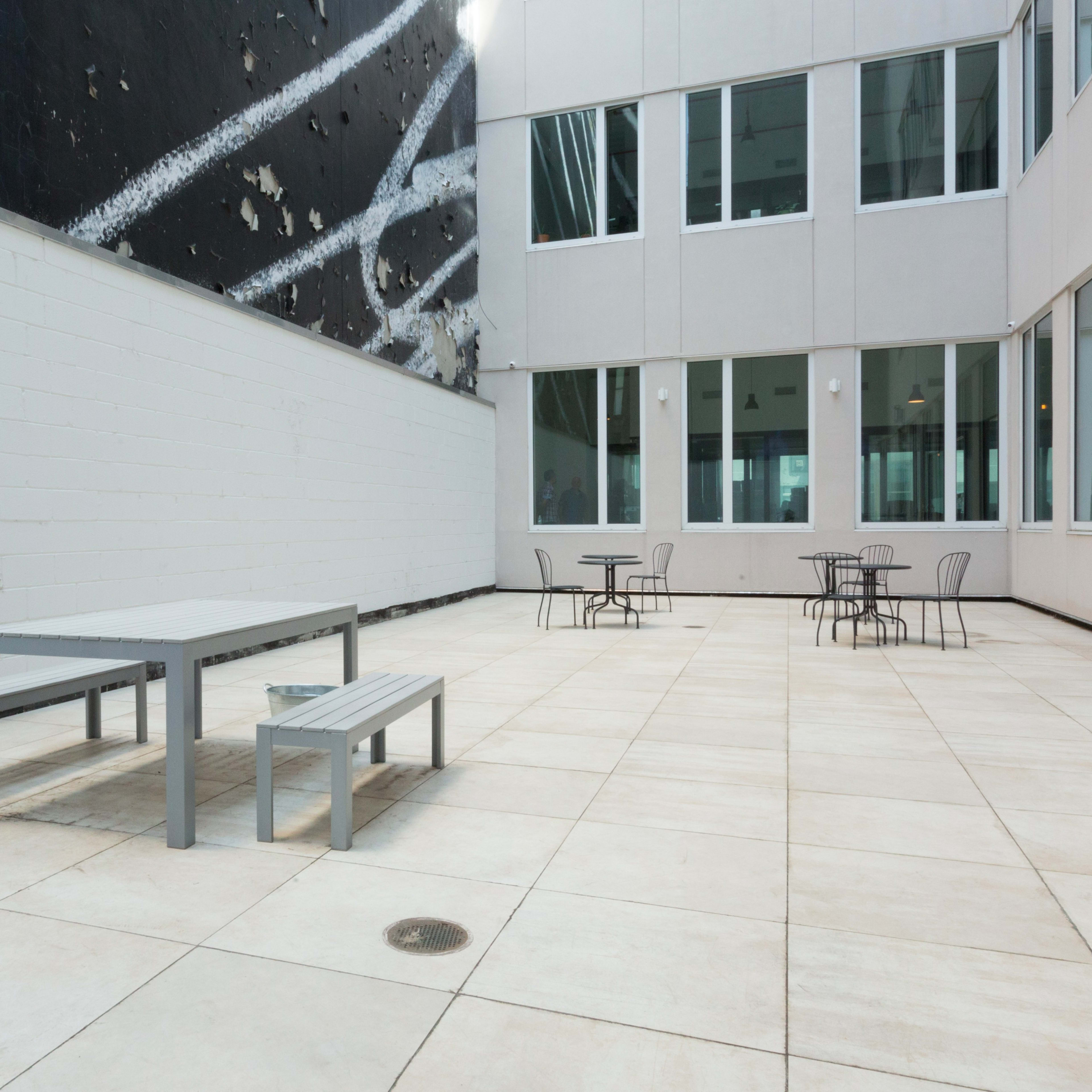 A modern outdoor courtyard with gray tables and benches surrounded by stark white walls and large windows.