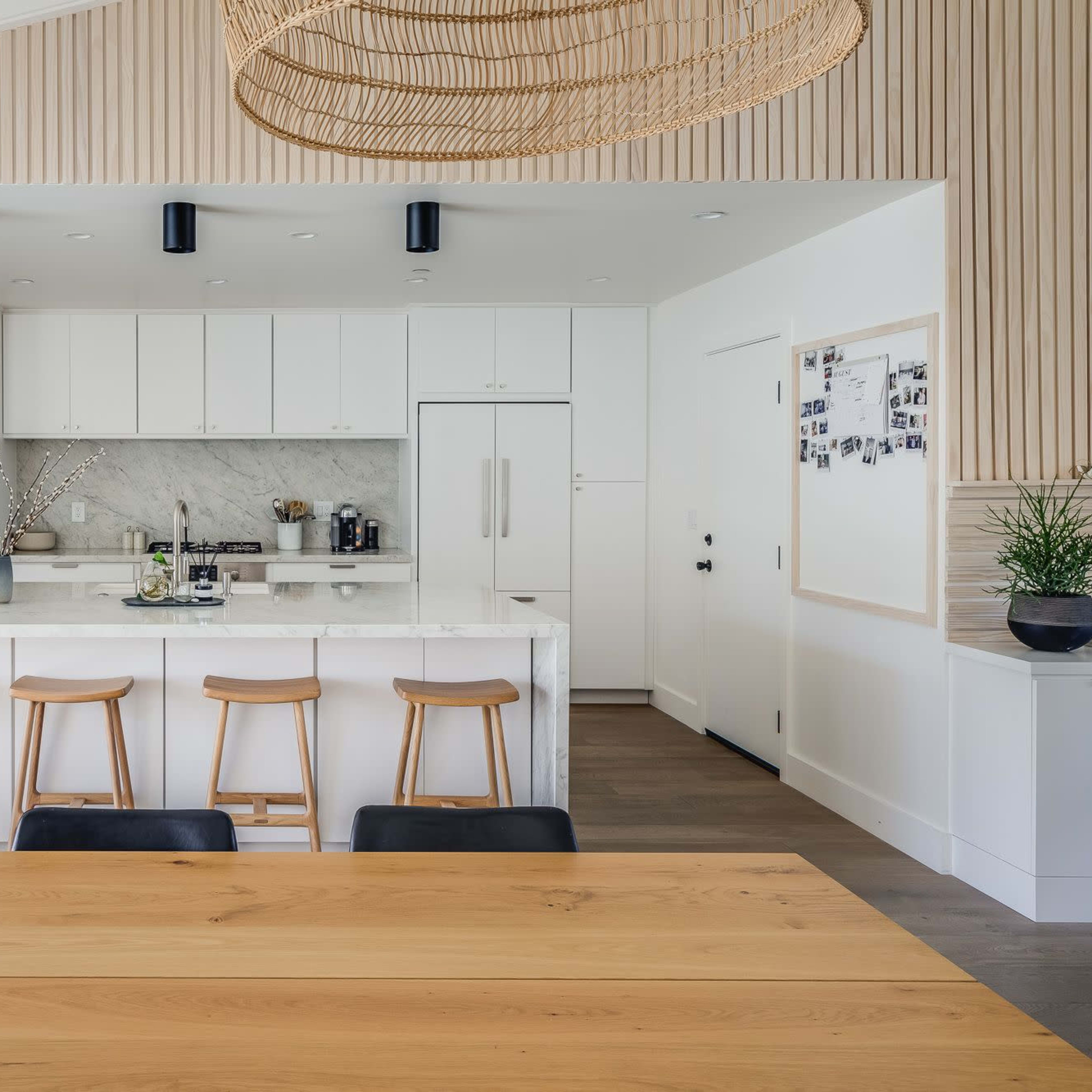 A modern kitchen features a marble island with four wooden stools, white cabinetry, and natural wood accents, adjacent to a dining table.