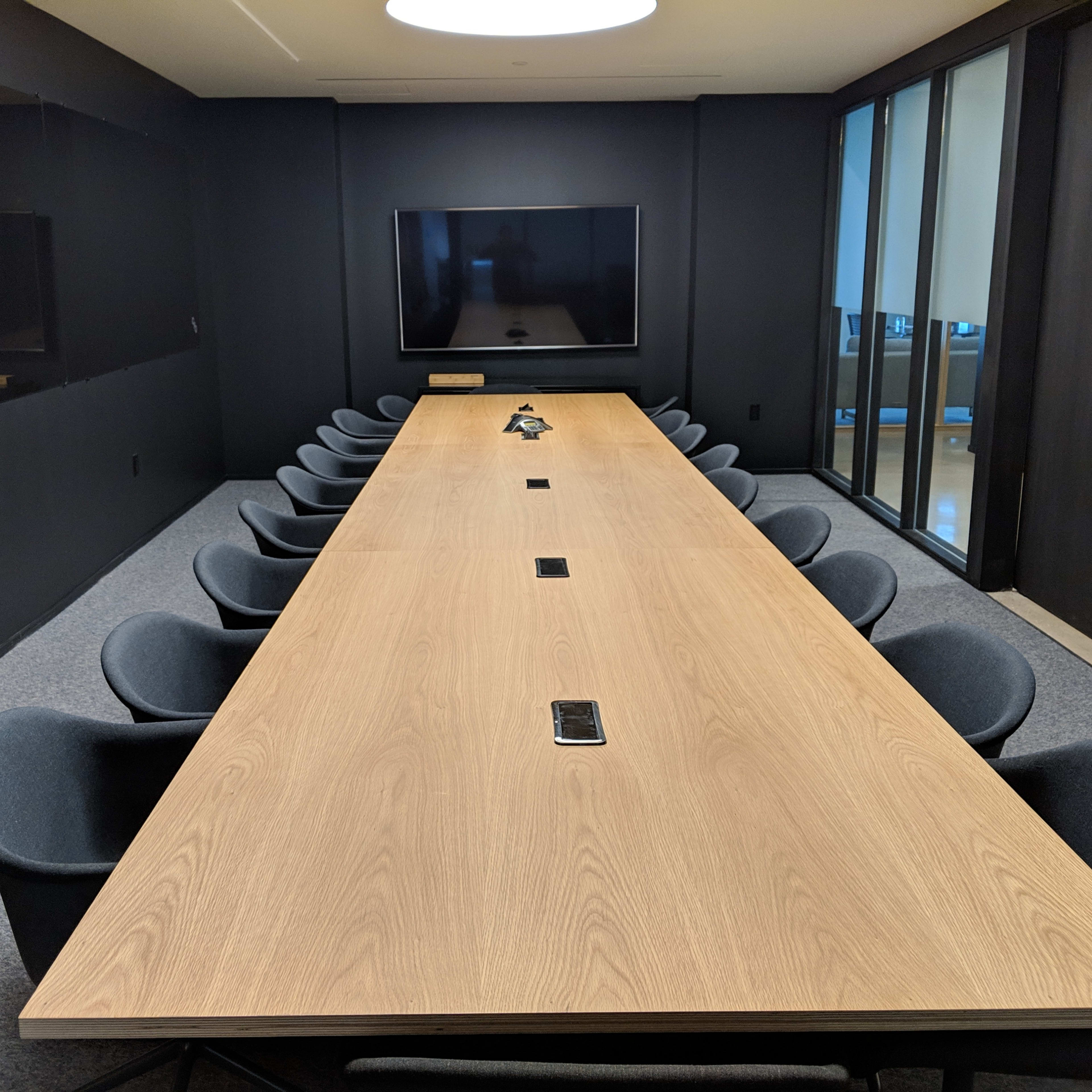 A large conference room with a rectangular wooden table surrounded by black chairs and a television mounted on the wall.
