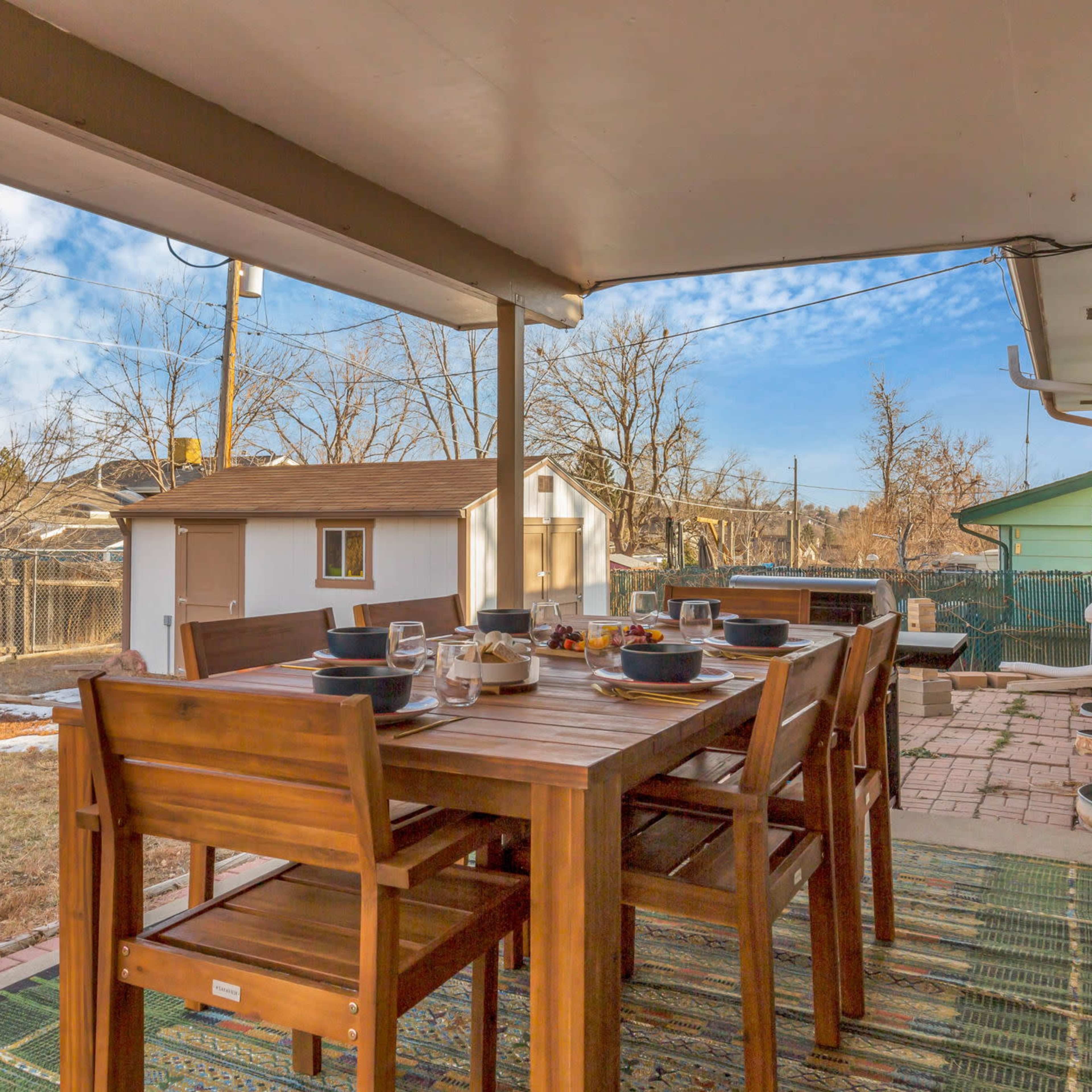 A wooden dining table is set for a meal on a patio with a view of a yard and nearby sheds under a partly cloudy sky.