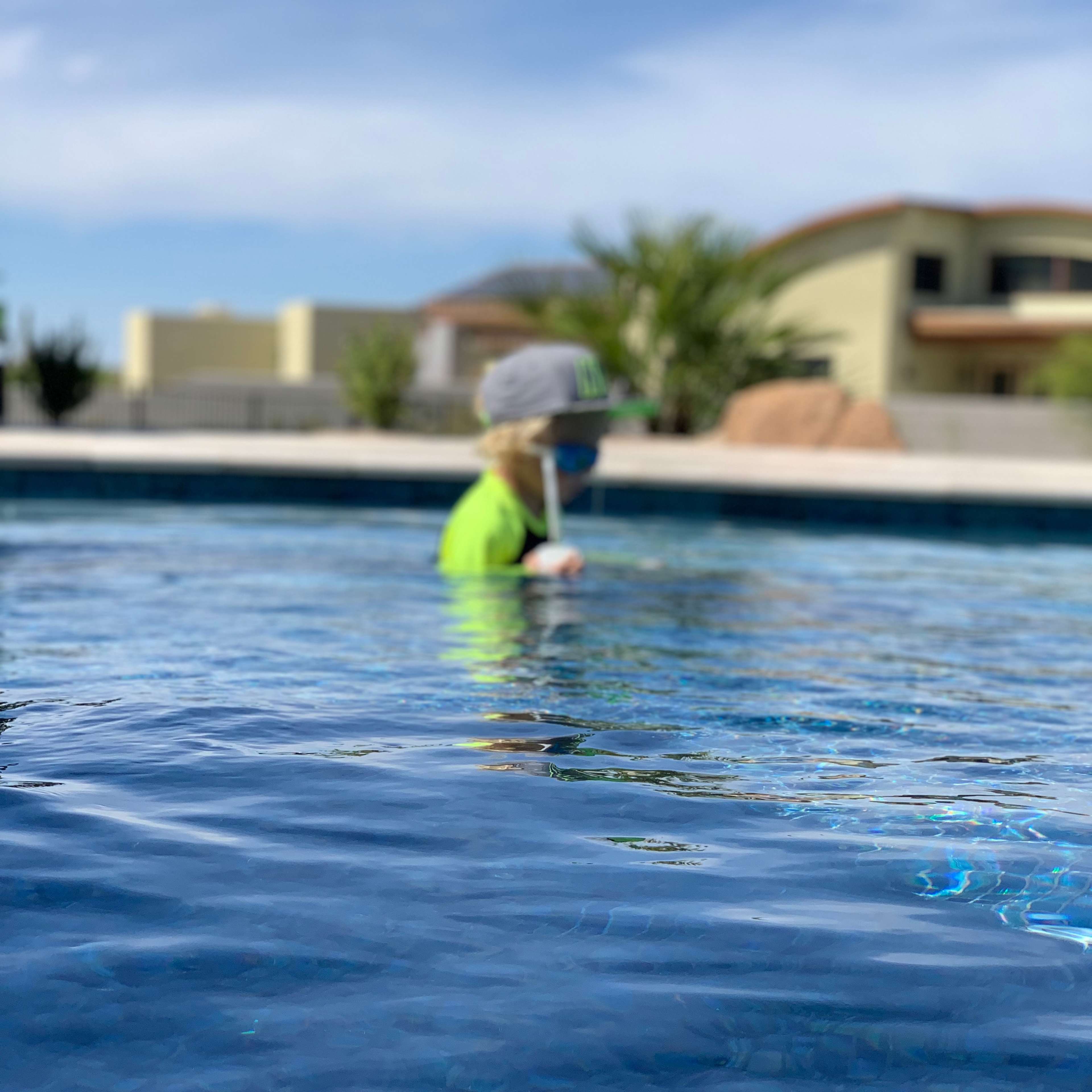A child in a green shirt and gray hat is standing in a swimming pool.