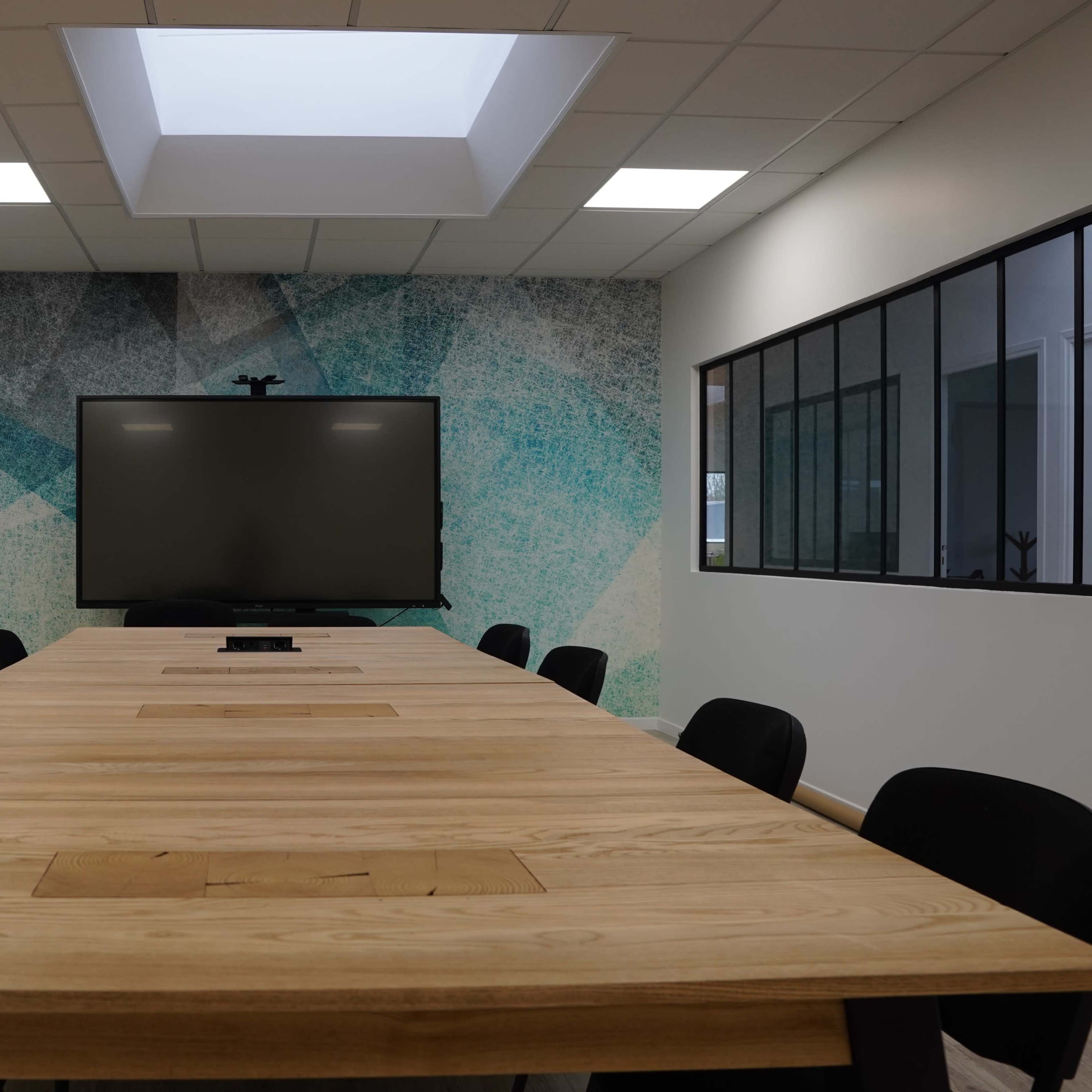 A modern conference room features a large wooden table surrounded by black chairs, with a wall-mounted screen and a skylight above.