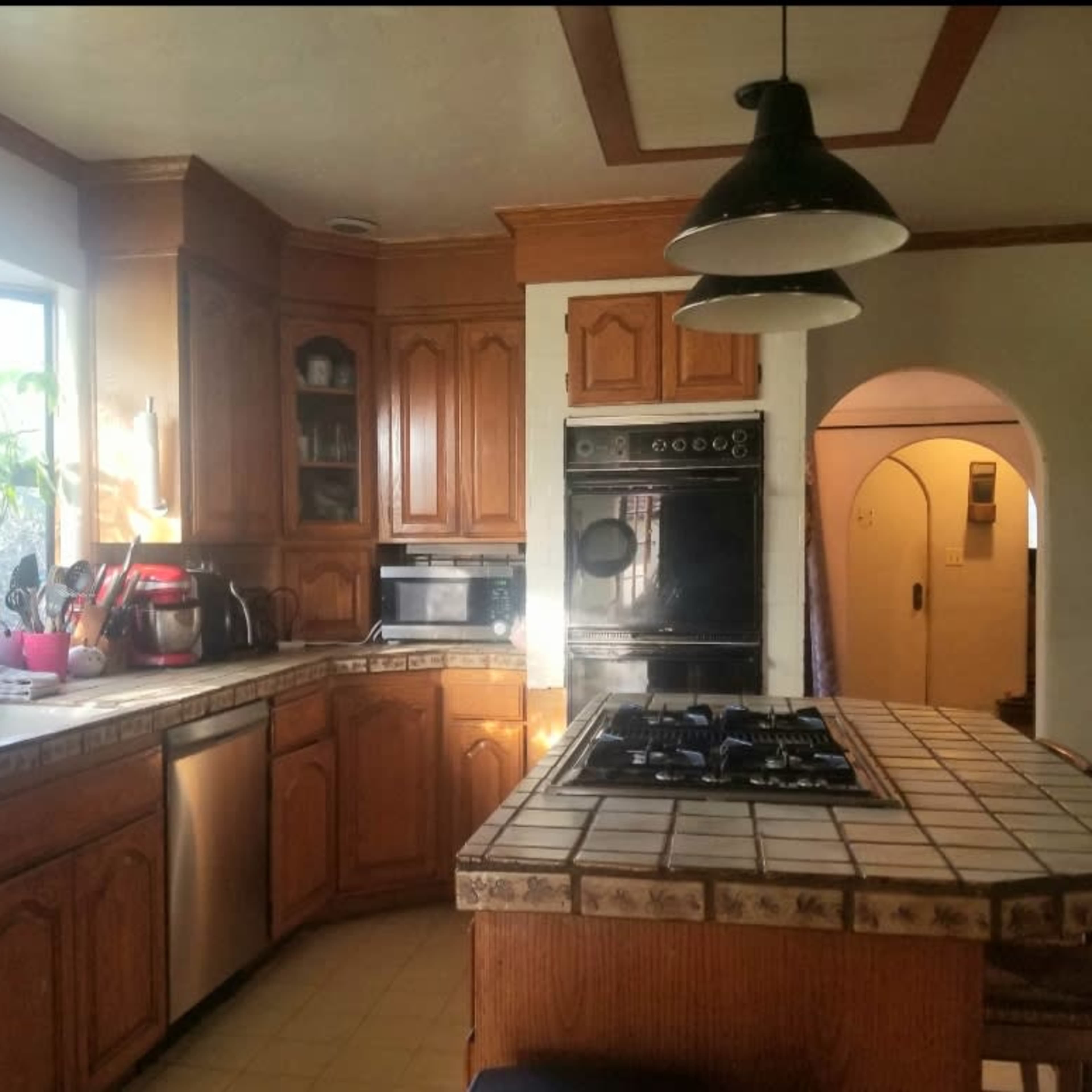 A kitchen features wooden cabinets, a tiled countertop, and various appliances including a stovetop, microwave, and sink.