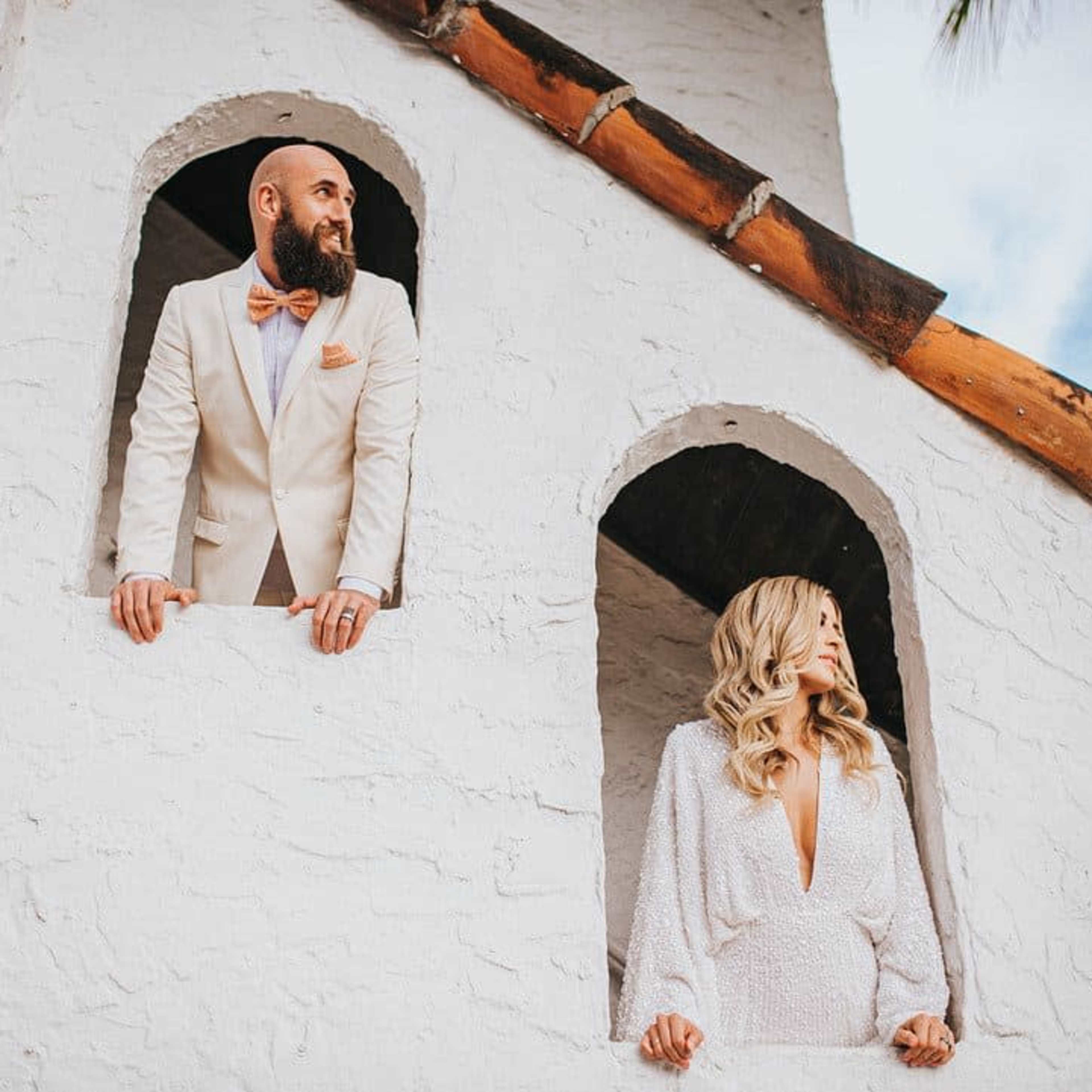 A man in a suit and a woman in a white dress stand in a building with arched openings.