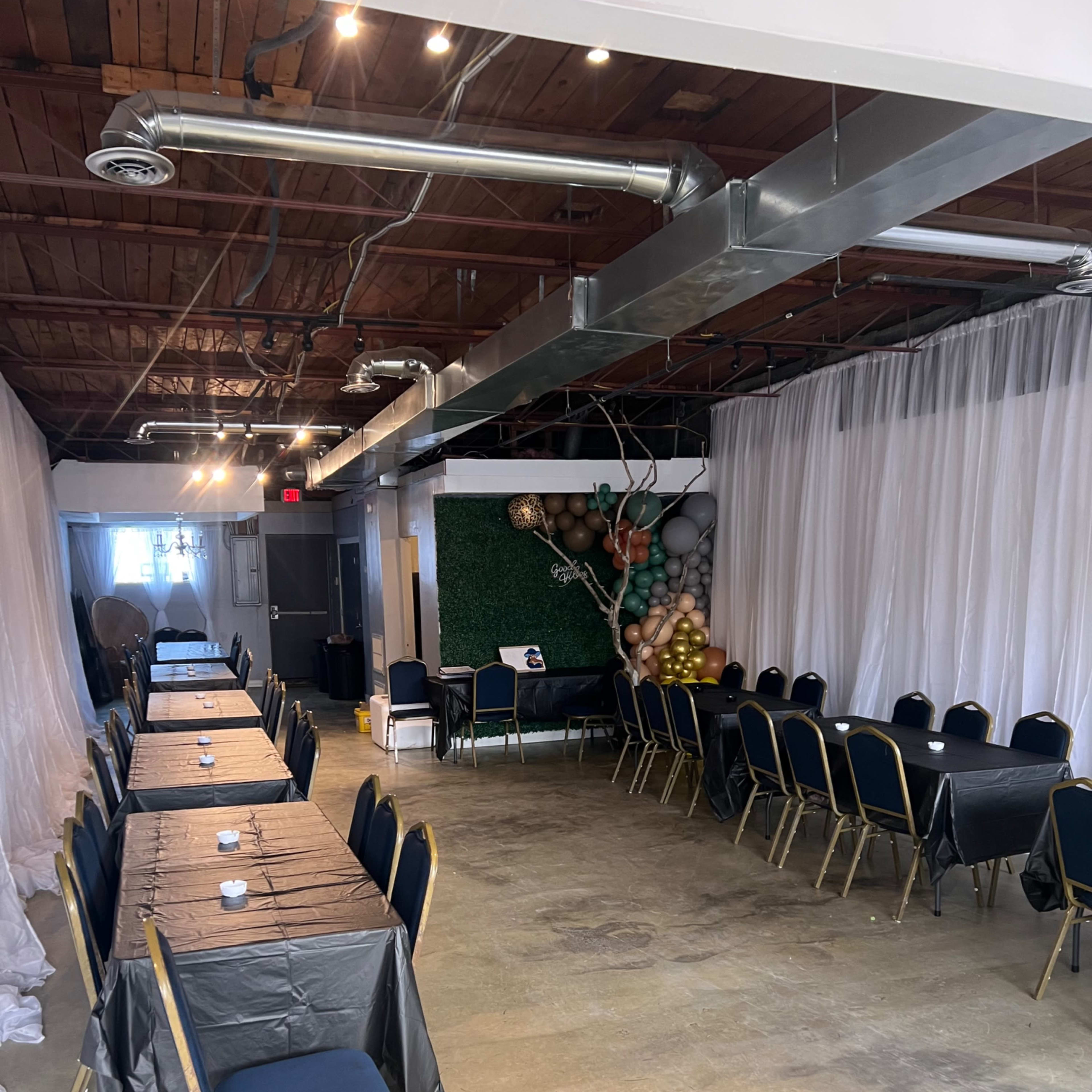 An empty event space with two long tables covered in black cloth, surrounded by blue chairs, and decorative elements including green foliage and balloons in one corner.