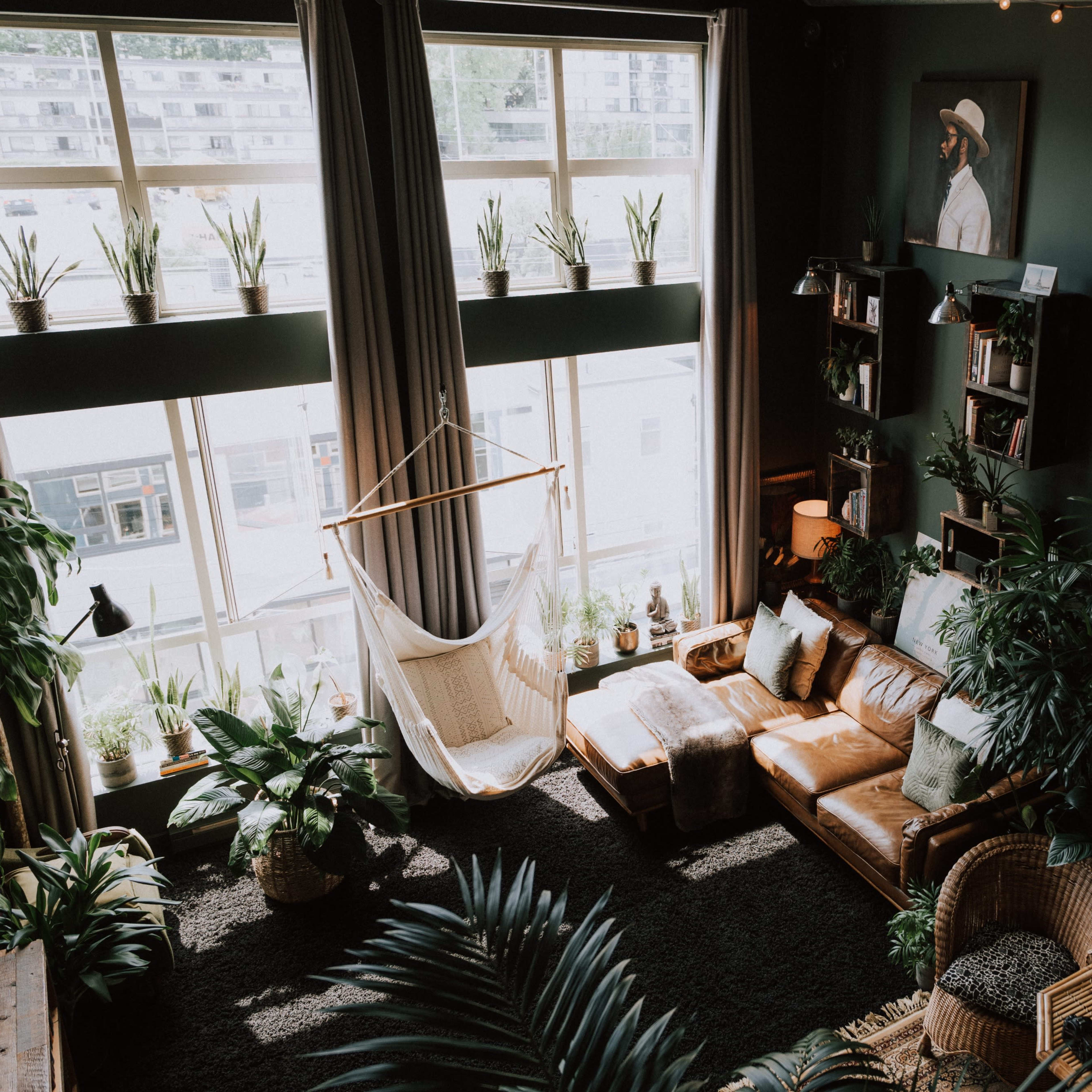 A spacious living room filled with various plants, featuring a large window, a hammock, leather couches, and a world map on the wall.