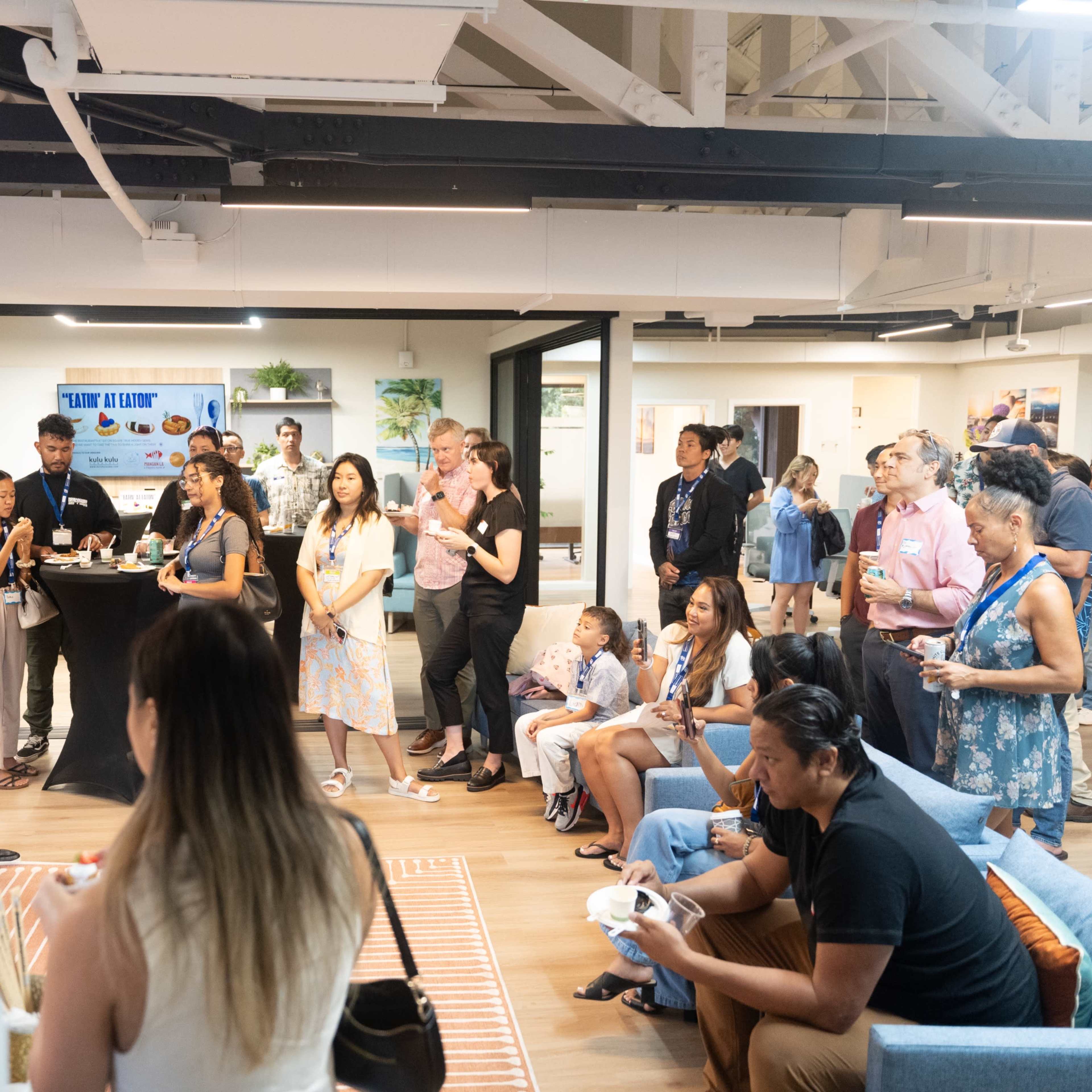 A diverse group of people gathers in a modern office space for a community event, with one man speaking at the front near a presentation display.