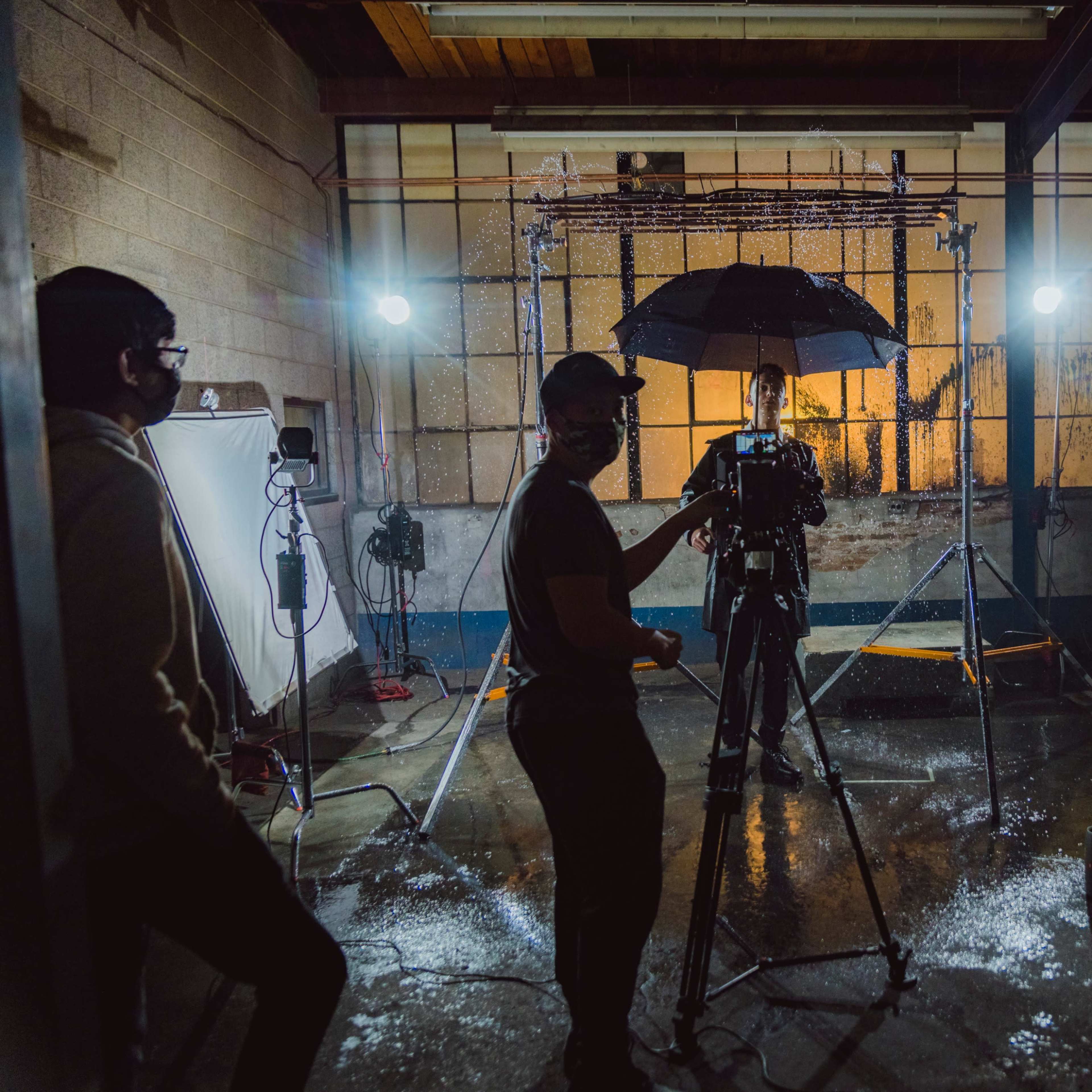 A film crew sets up equipment in a dimly lit, industrial space with a model holding an umbrella under studio lights.
