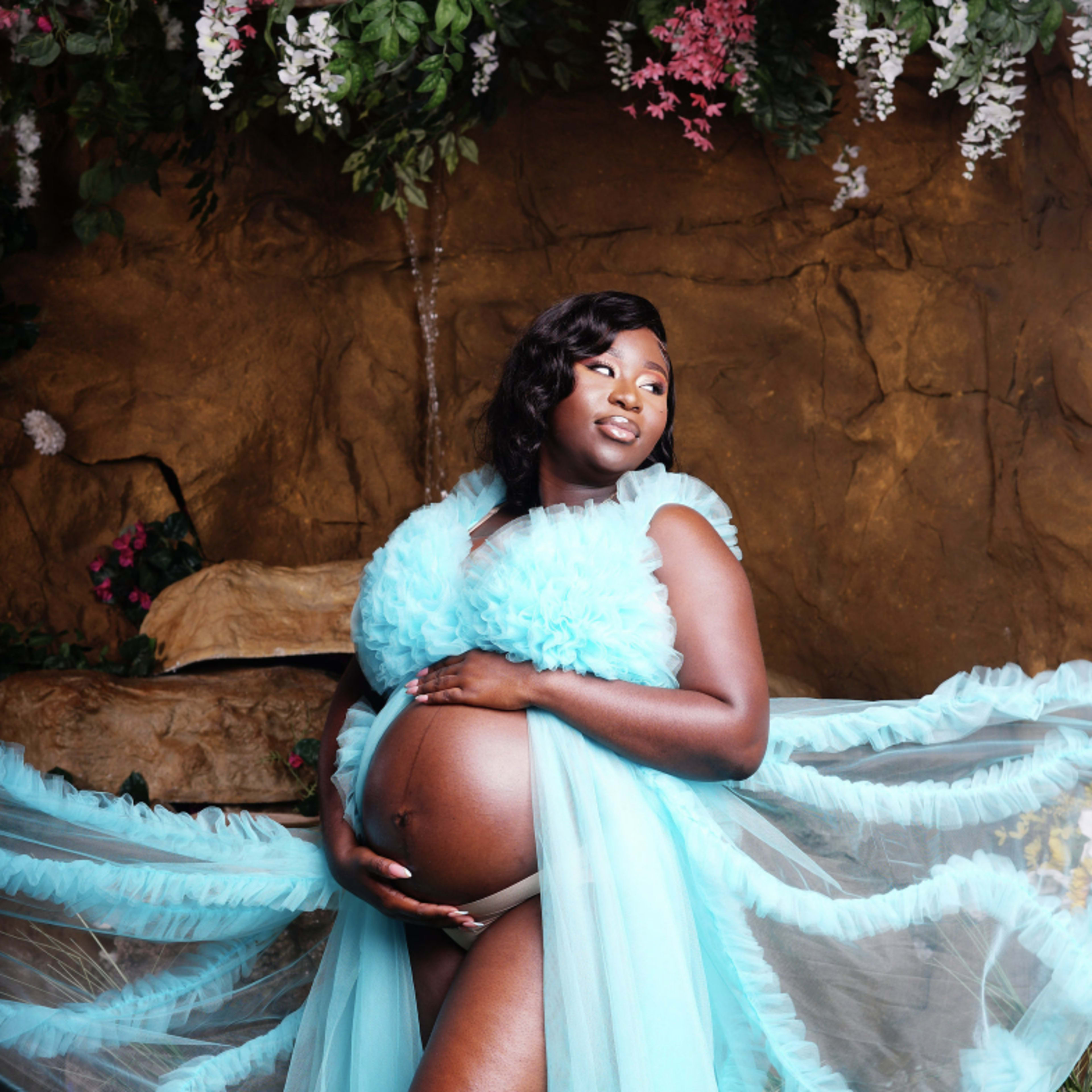 A pregnant woman poses with her hands on her belly, wearing a flowing, sheer blue garment in a floral setting.