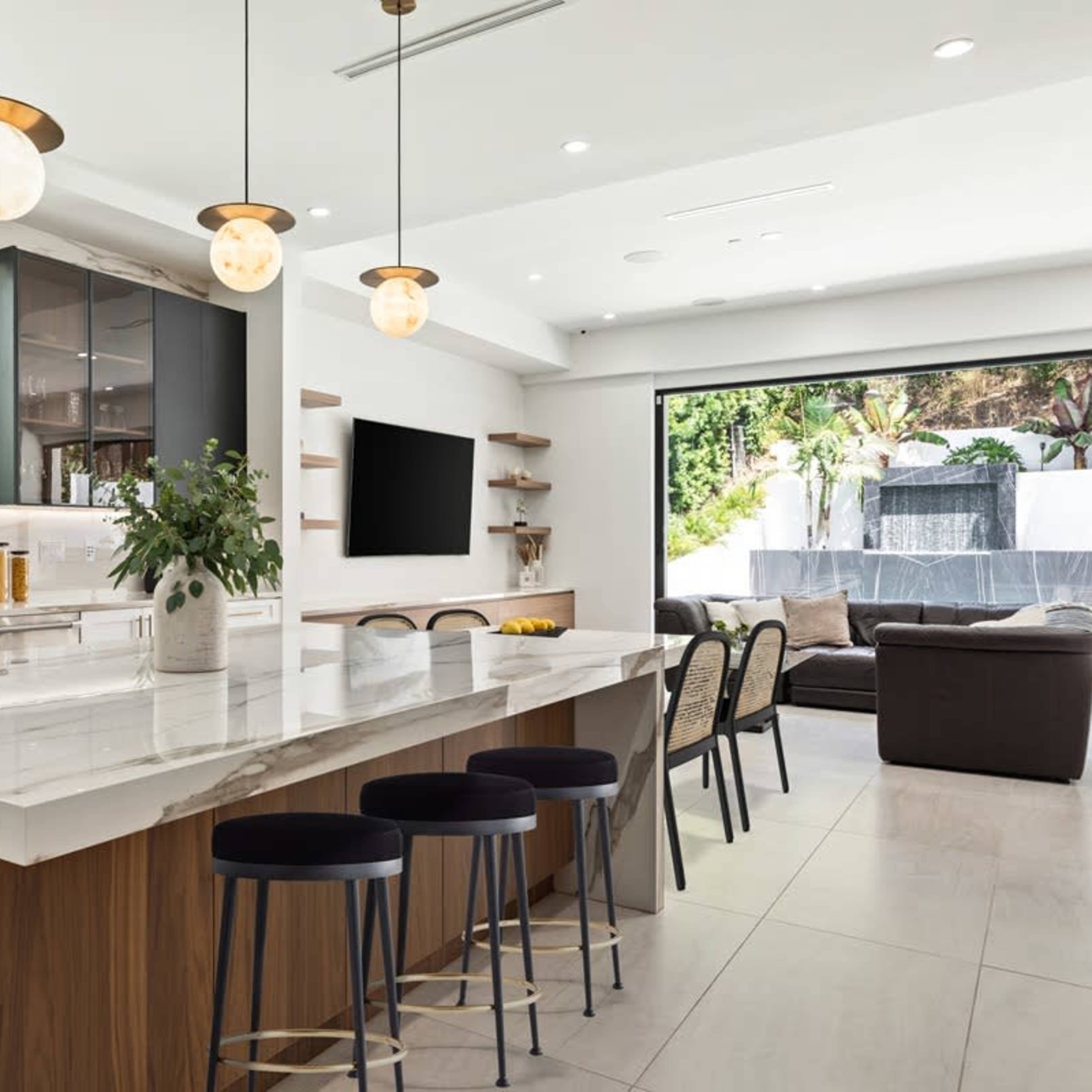 A modern kitchen with a large island, contemporary bar stools, and a glass door leading to an outdoor seating area.