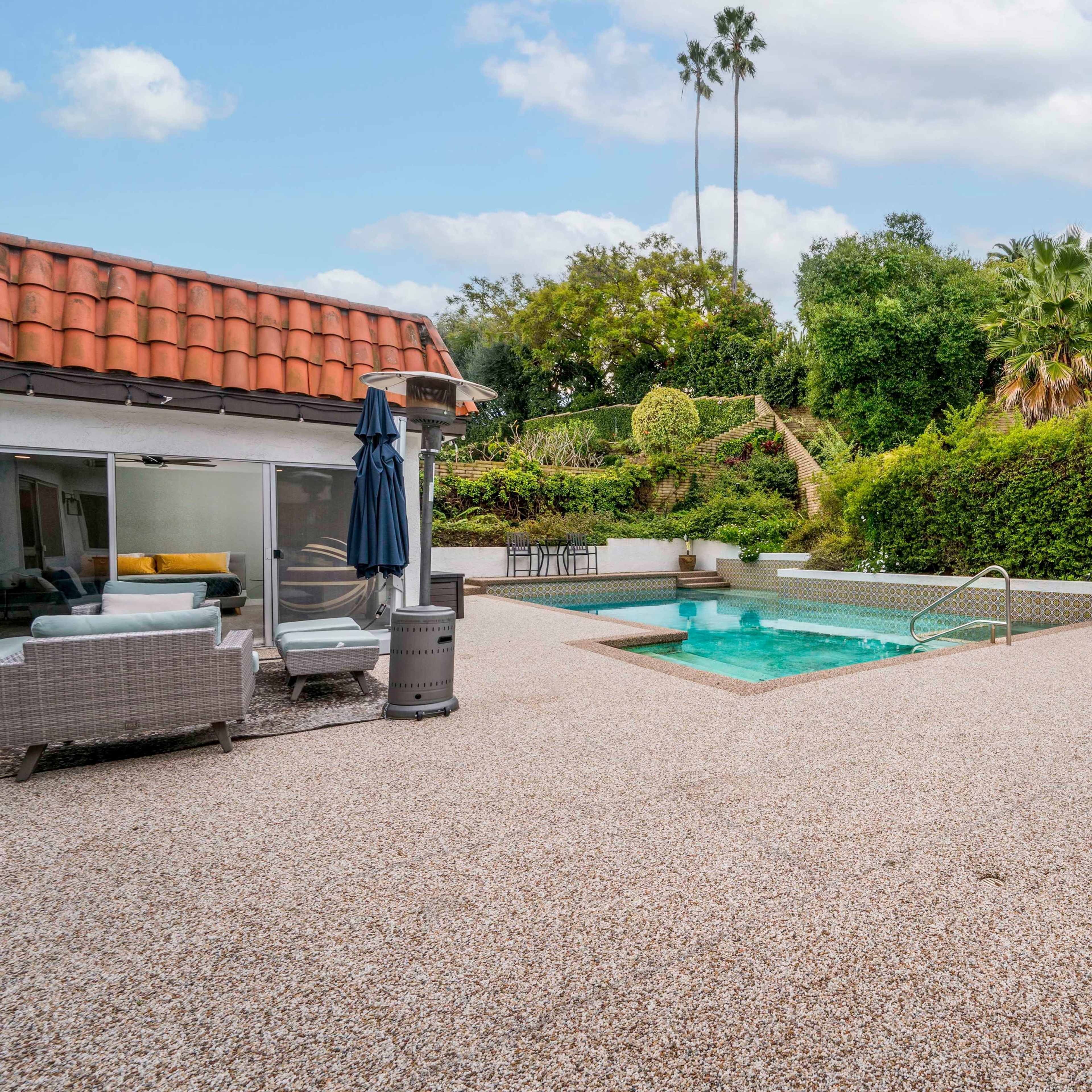 A backyard with a pool surrounded by a gravel patio, lounge chairs, and lush greenery in the background.