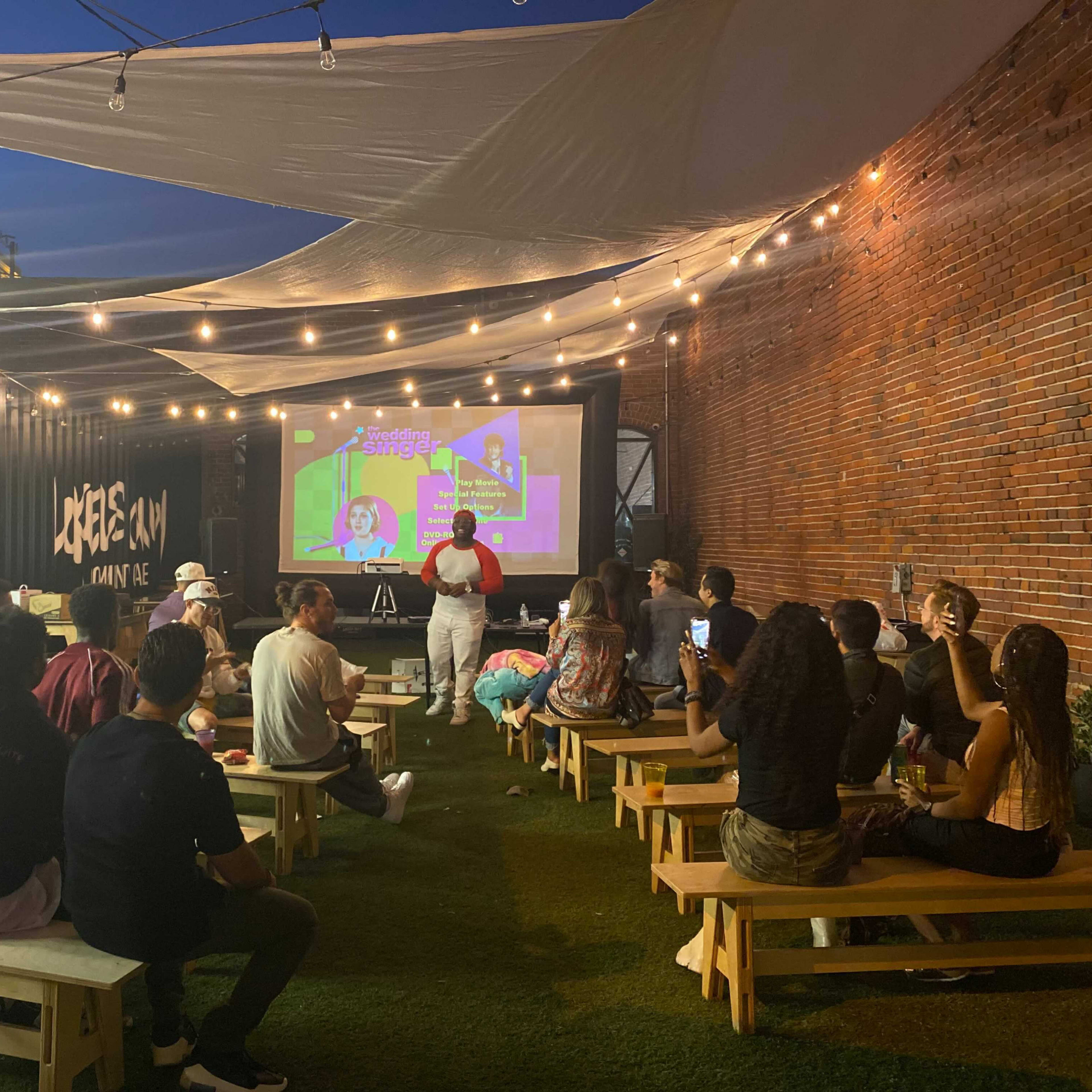 A group of people are seated on benches in an outdoor space, watching a presentation on a large screen illuminated by string lights.