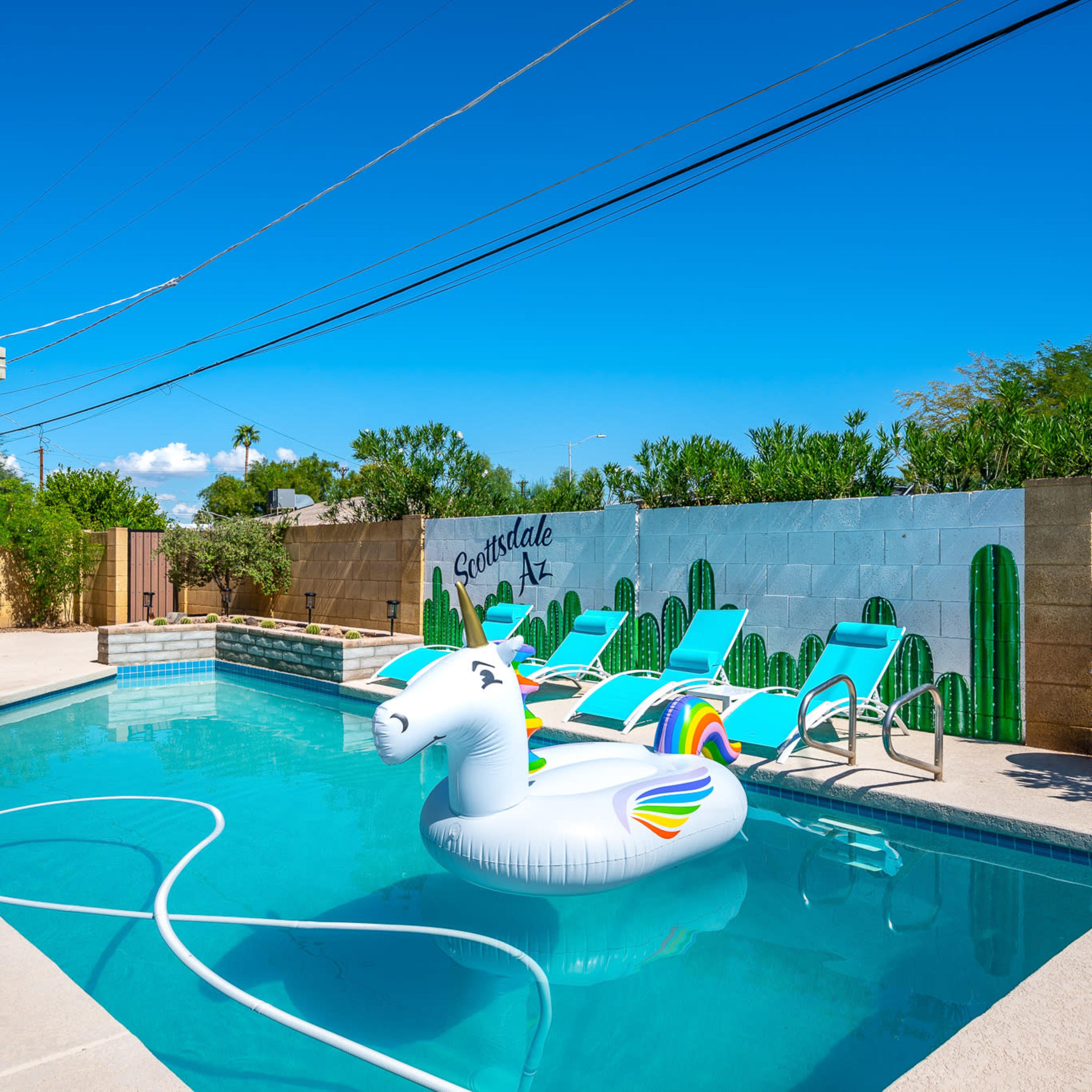 A swimming pool with a unicorn float and lounge chairs is surrounded by a mural of cacti against a clear blue sky.
