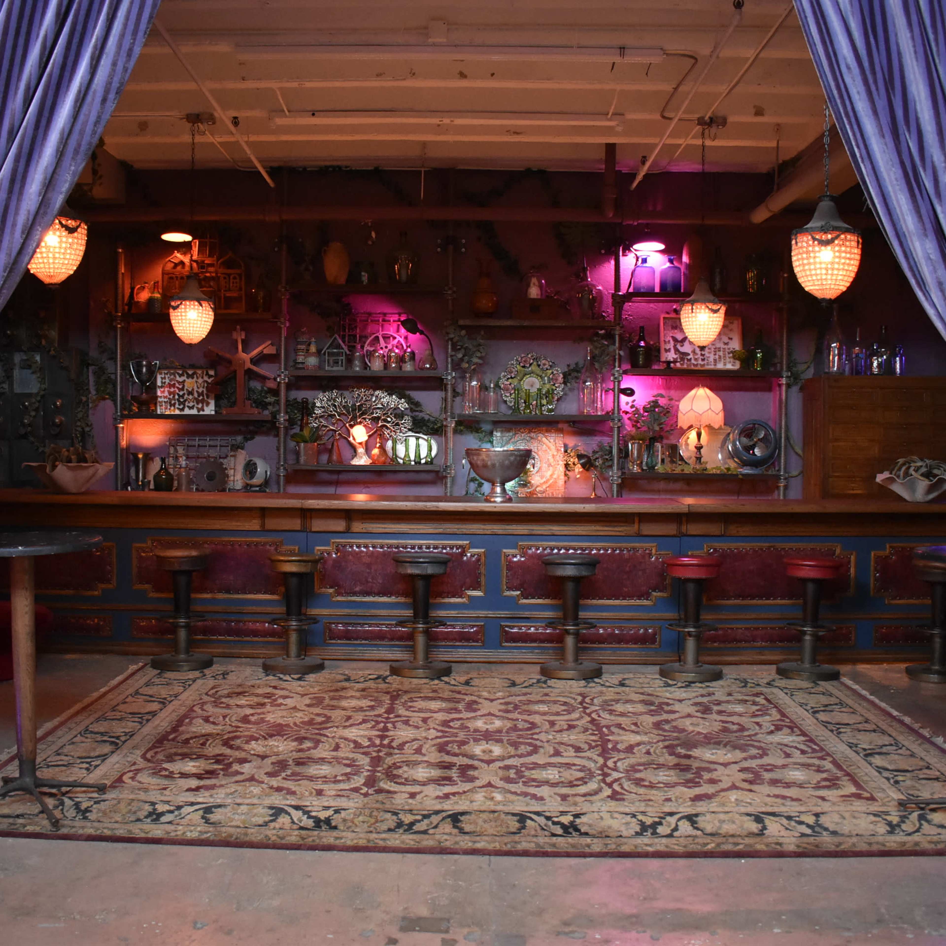 A dimly lit bar area with high stools, a rug on the floor, and shelves adorned with various decorative items, flanked by long, purple curtains.