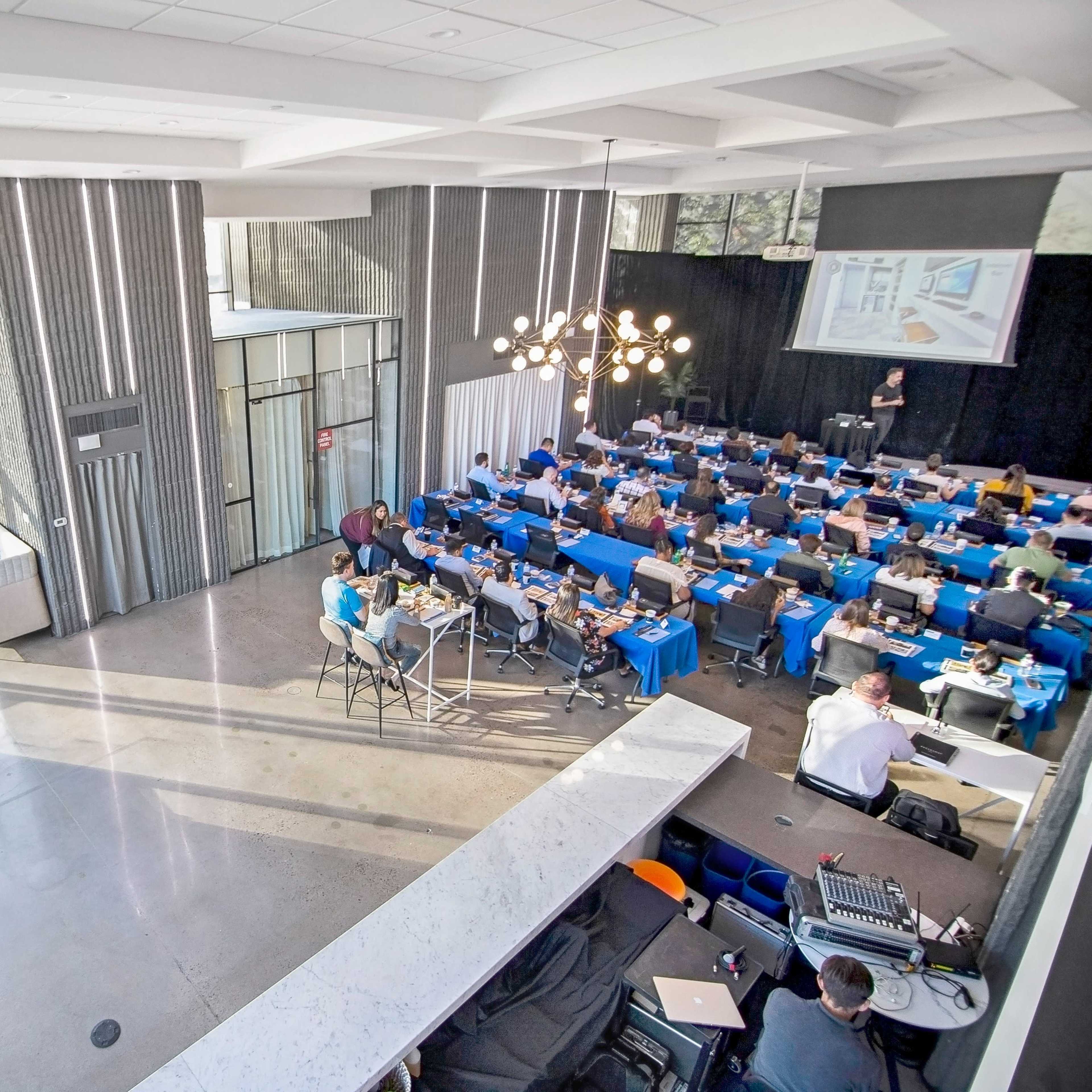 A well-attended conference room with rows of tables set up for participants, including equipment and refreshments, while a speaker presents at the front.