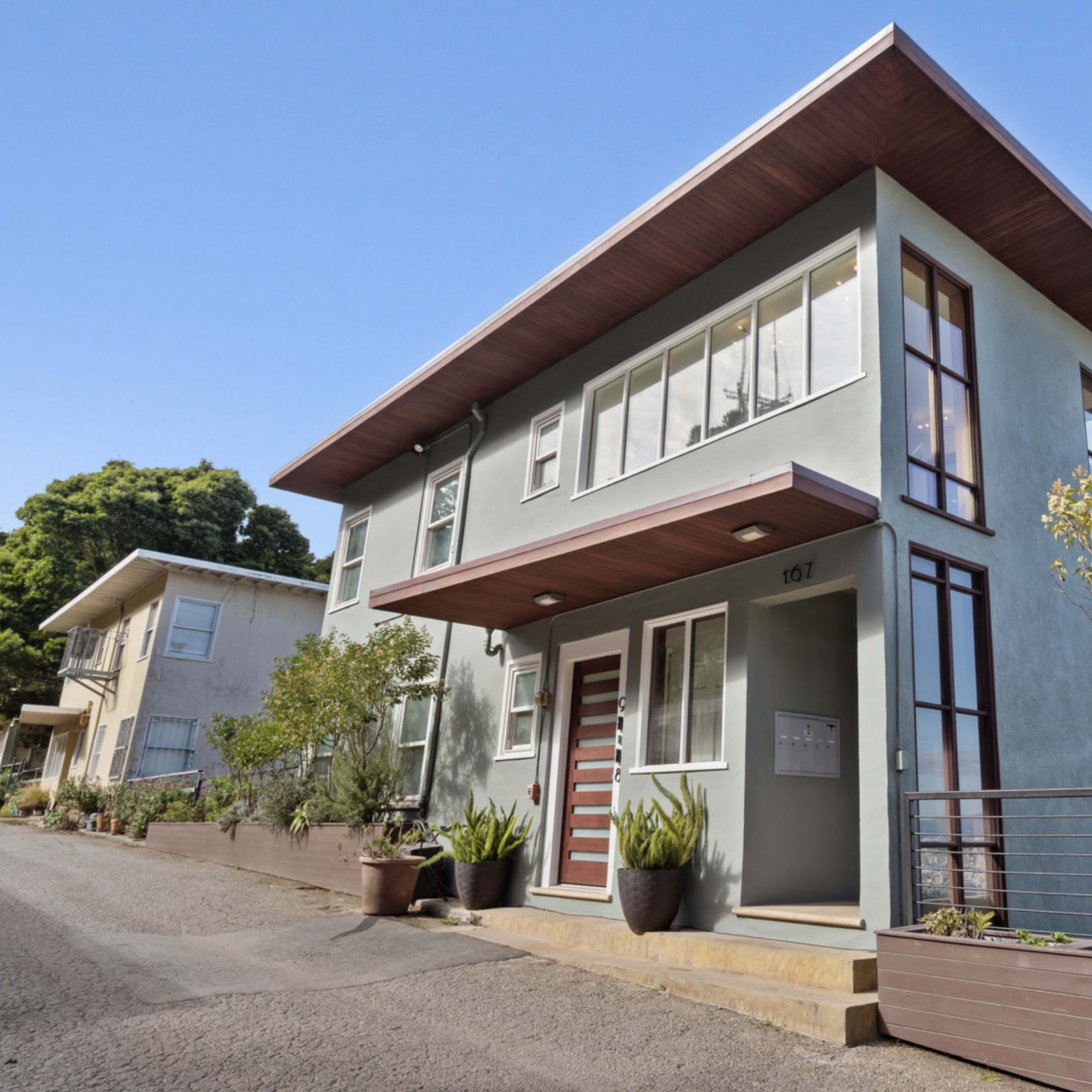 A modern two-story house with large windows, a flat roof, and a landscaped front area is situated on a sloped street.