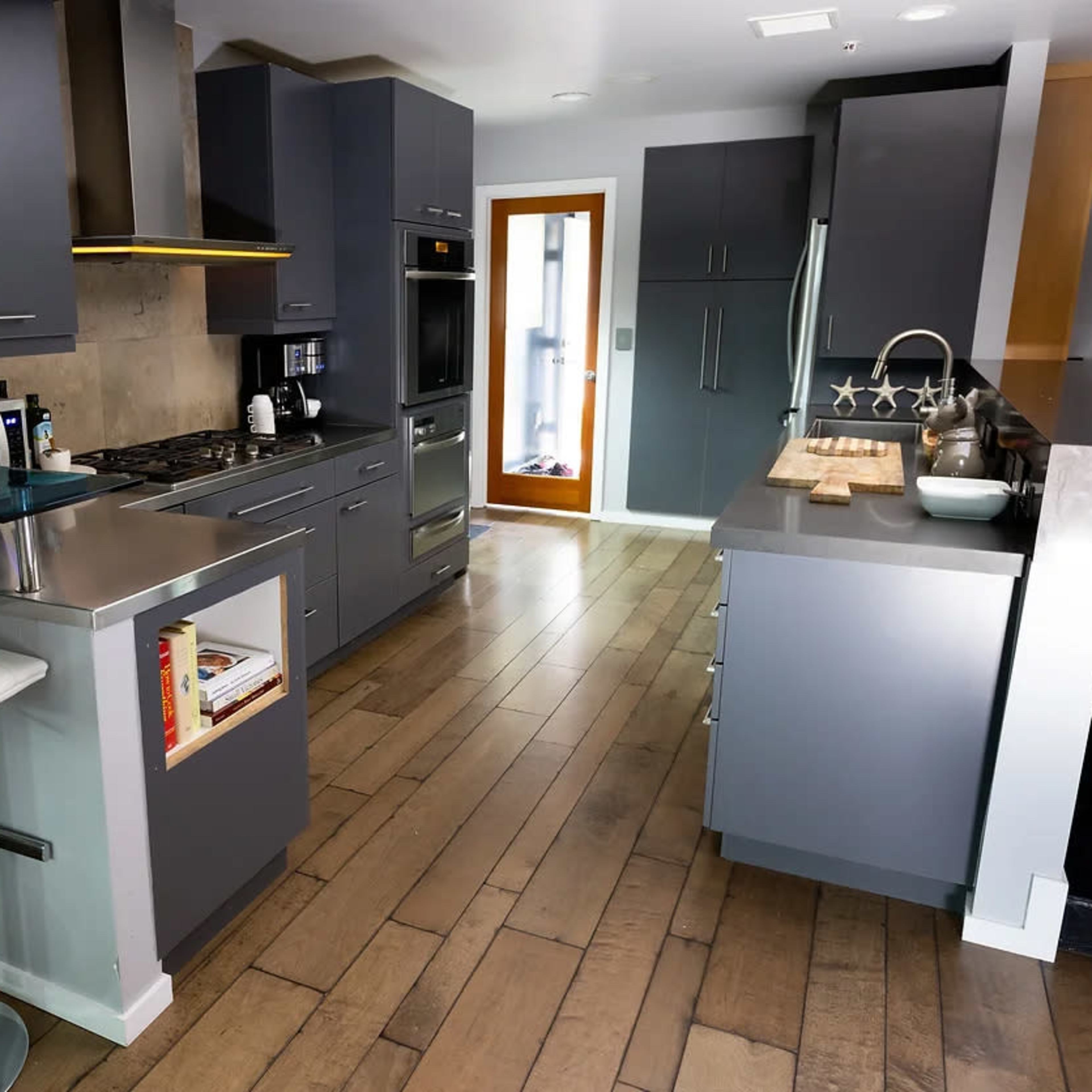 A modern kitchen with gray cabinets, stainless steel appliances, a bar with two white stools, and a wooden floor.