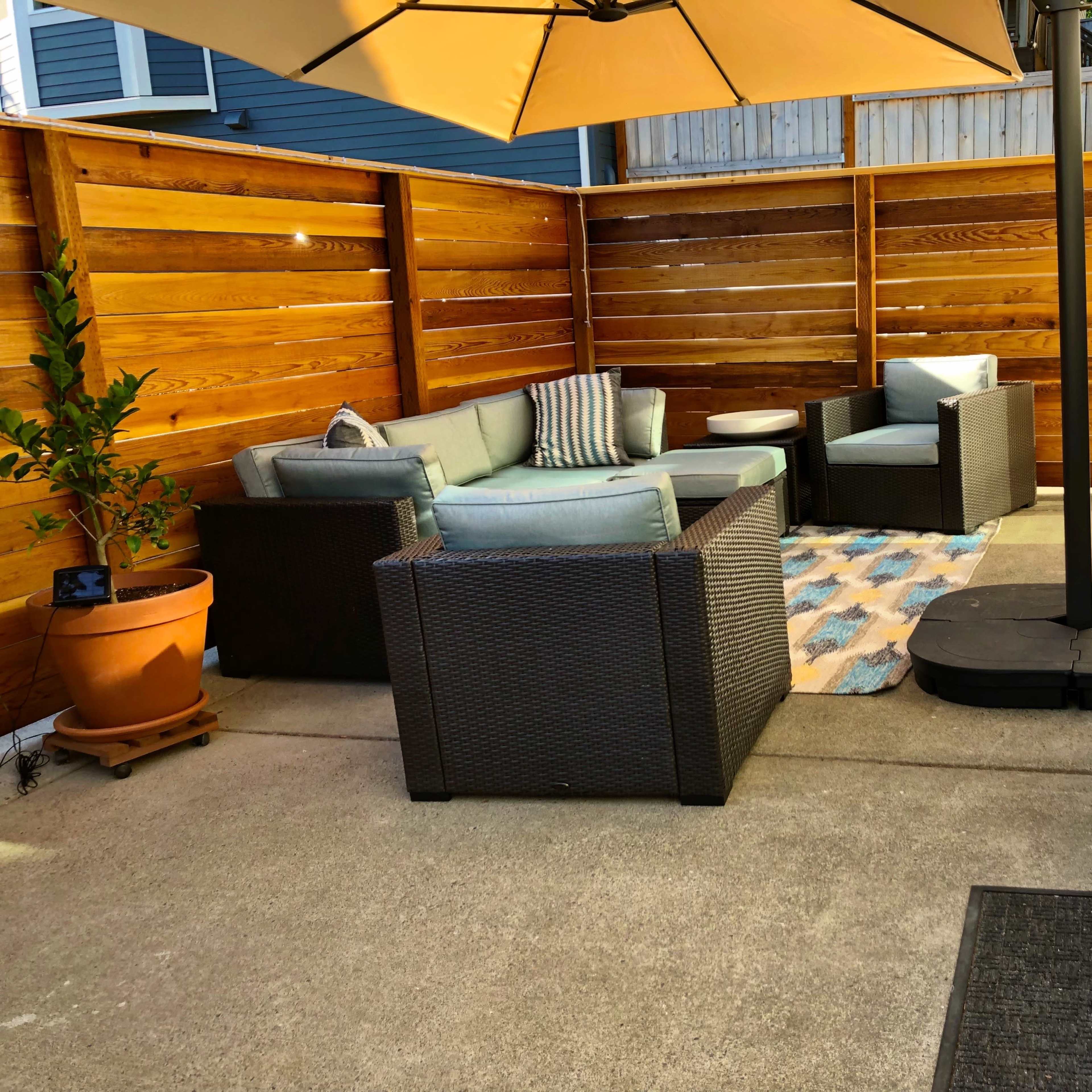 A cozy outdoor seating area features a sofa set, coffee table, and umbrella on a concrete patio surrounded by wooden fencing.