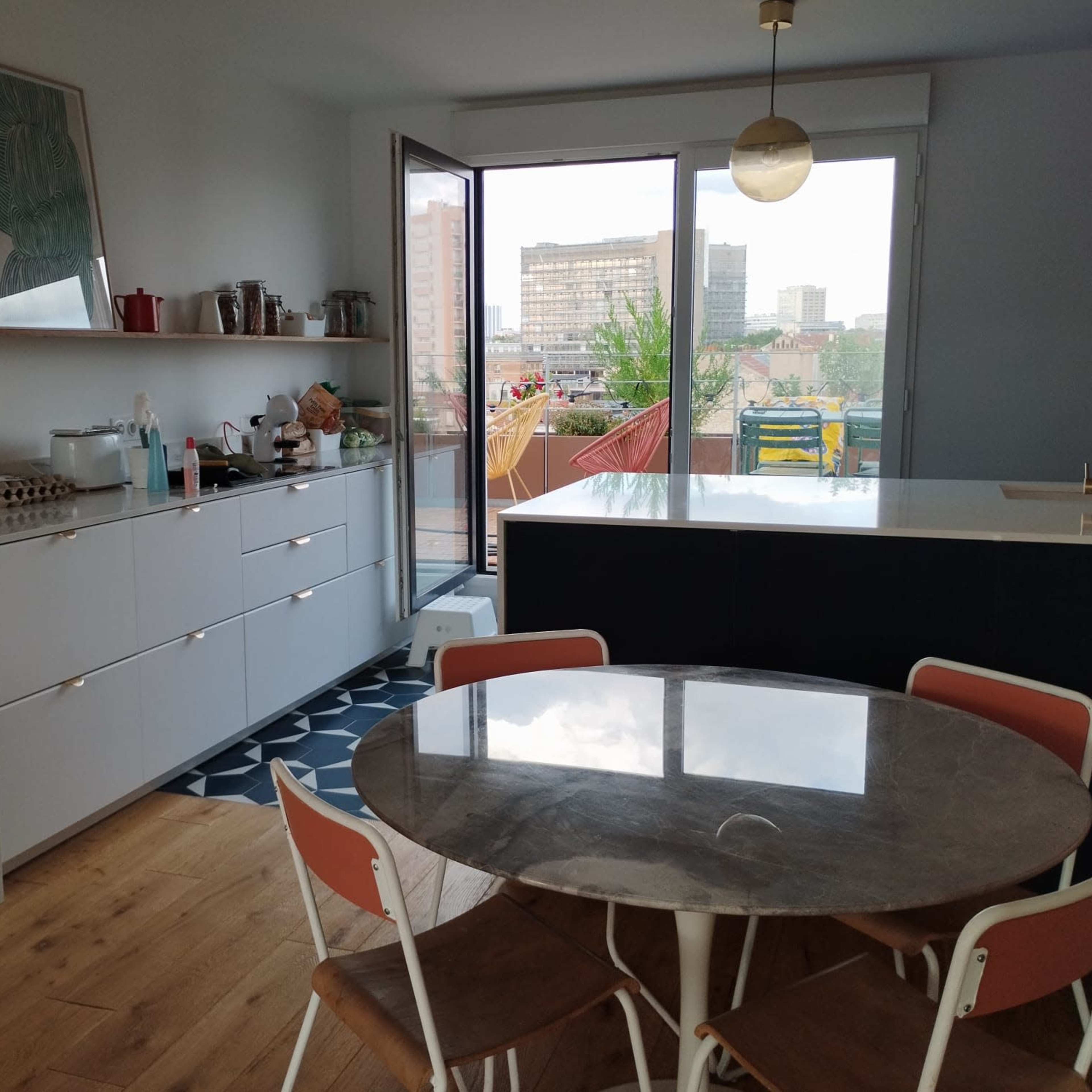 A modern kitchen features a round table with white chairs, a marble-topped counter, and a large window overlooking a cityscape.