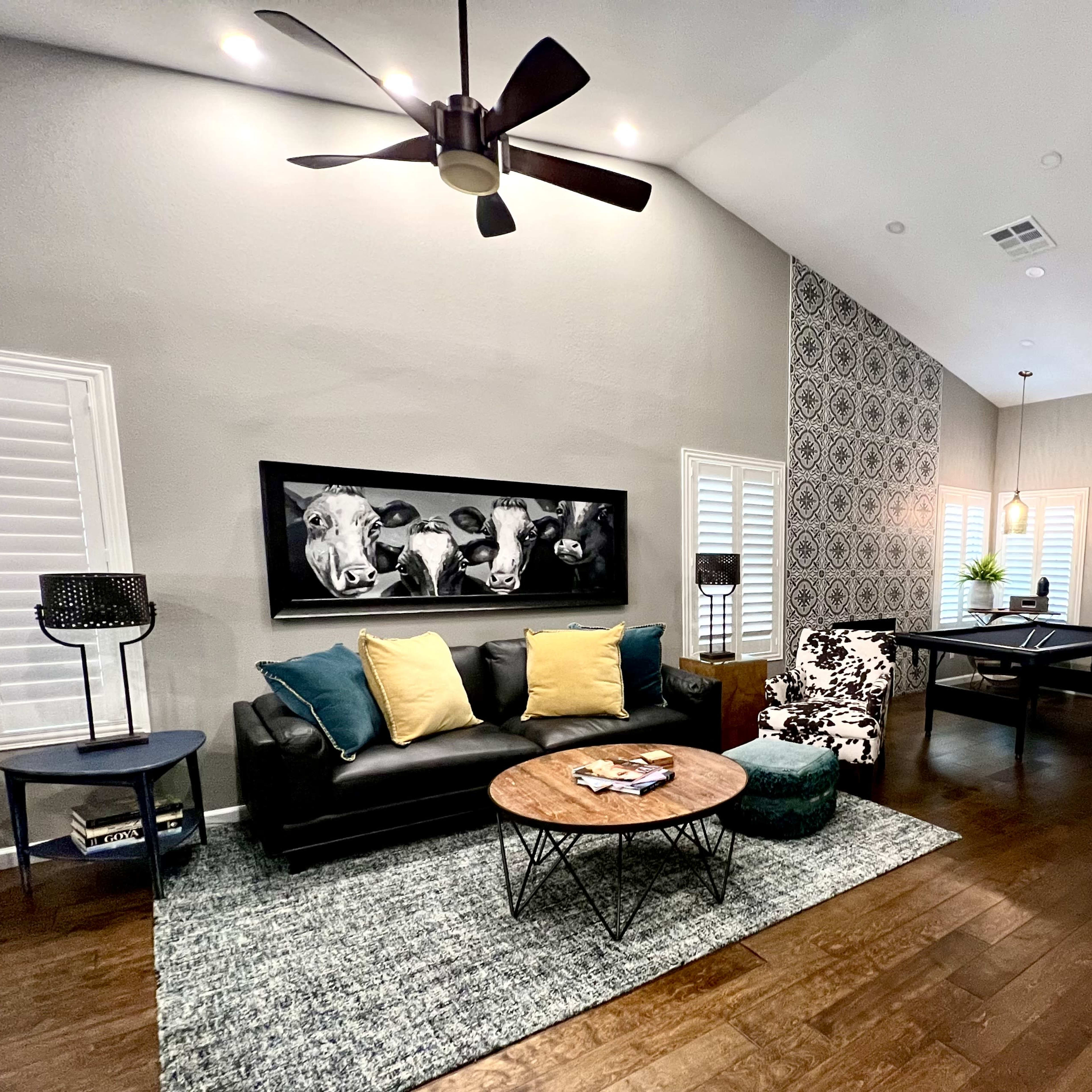 A modern living room features a black leather sofa with yellow cushions, a round coffee table, and a billiard table in the background.