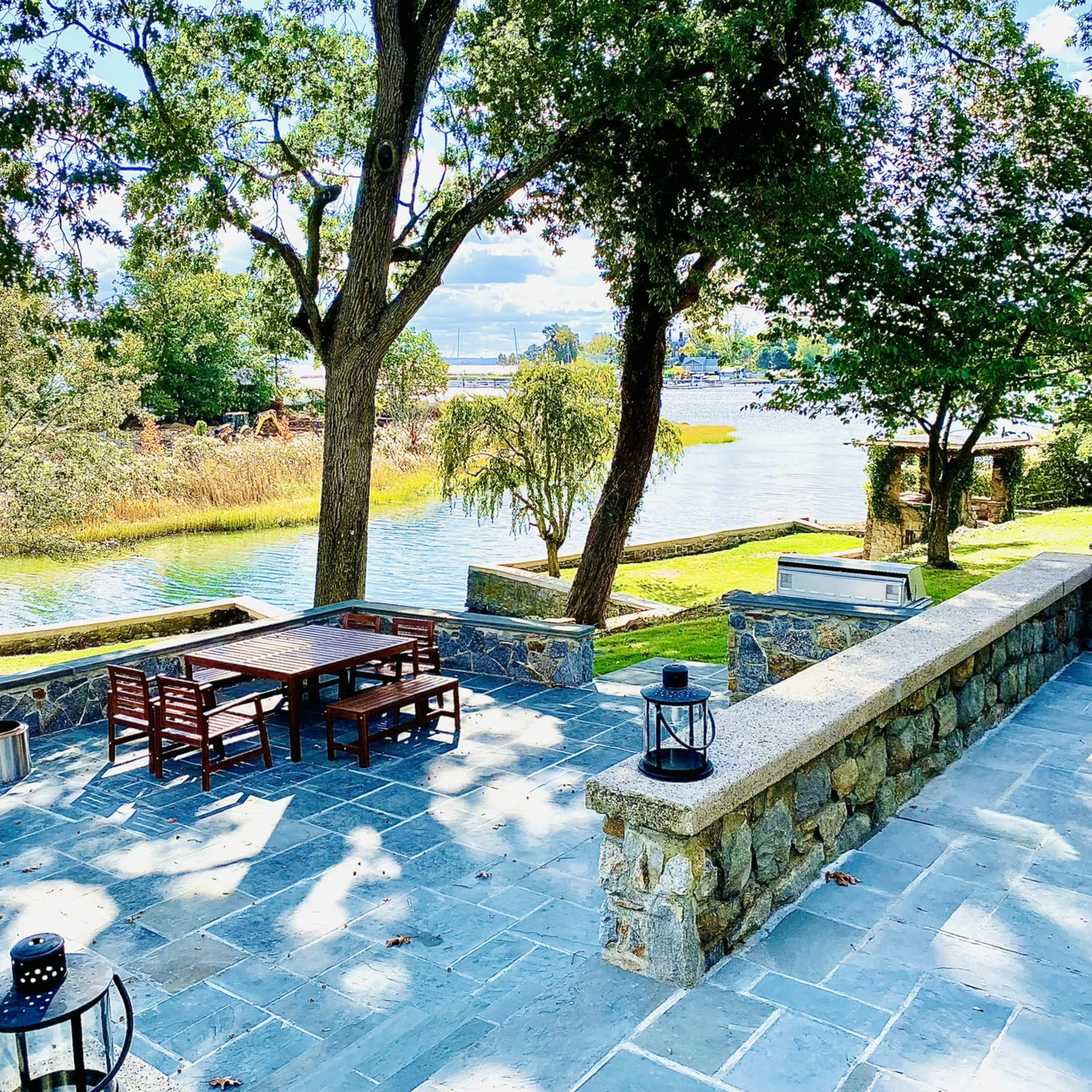 The scene shows a stone patio with wooden tables and chairs, overlooking a river surrounded by trees and grass.