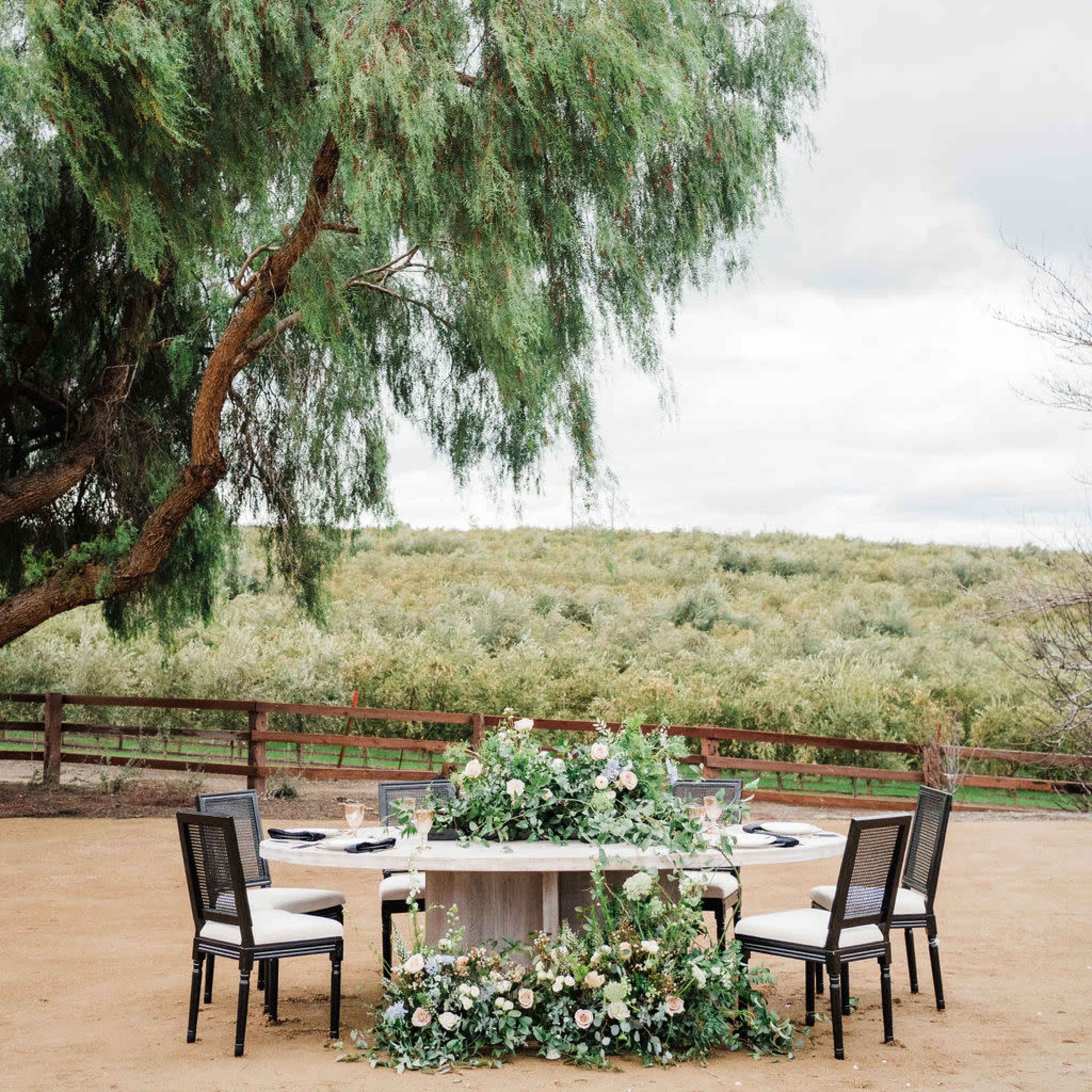A dining table adorned with floral arrangements is set outdoors under a large tree, surrounded by chairs and a scenic landscape in the background.