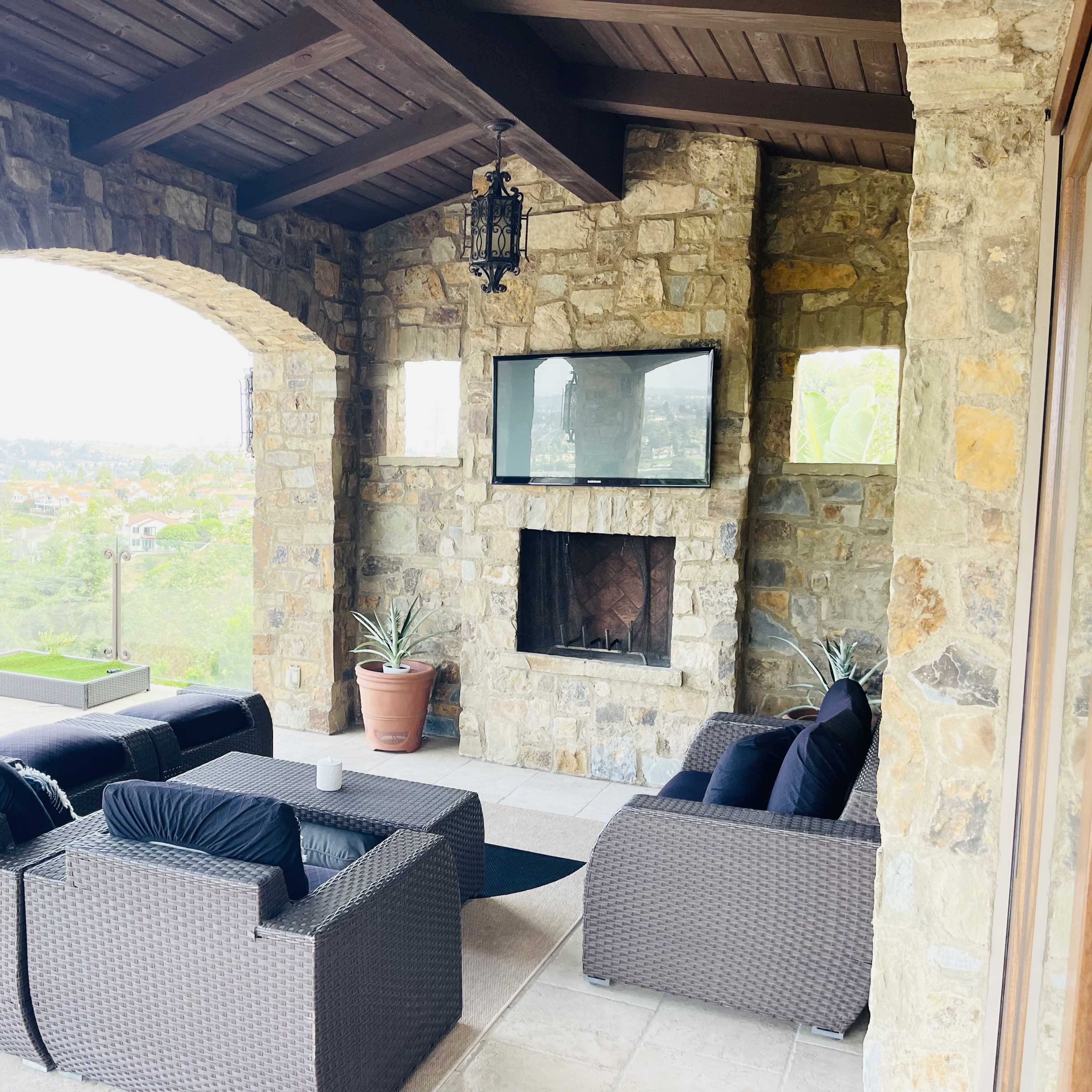 A stone patio with a seating area featuring dark wicker furniture and a fireplace, overlooking a scenic view.