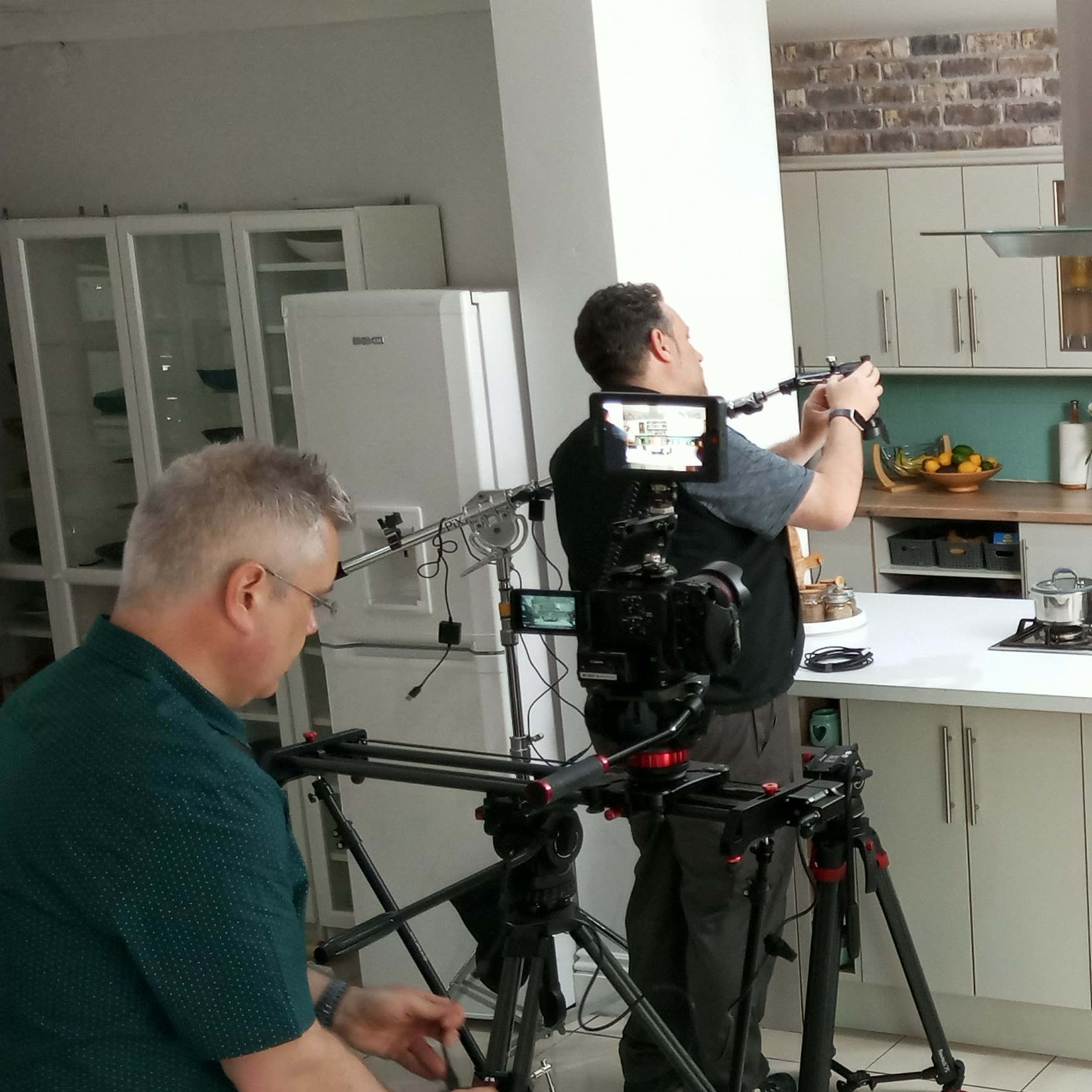 A film crew sets up equipment in a modern kitchen while one person prepares food and another operates a camera.