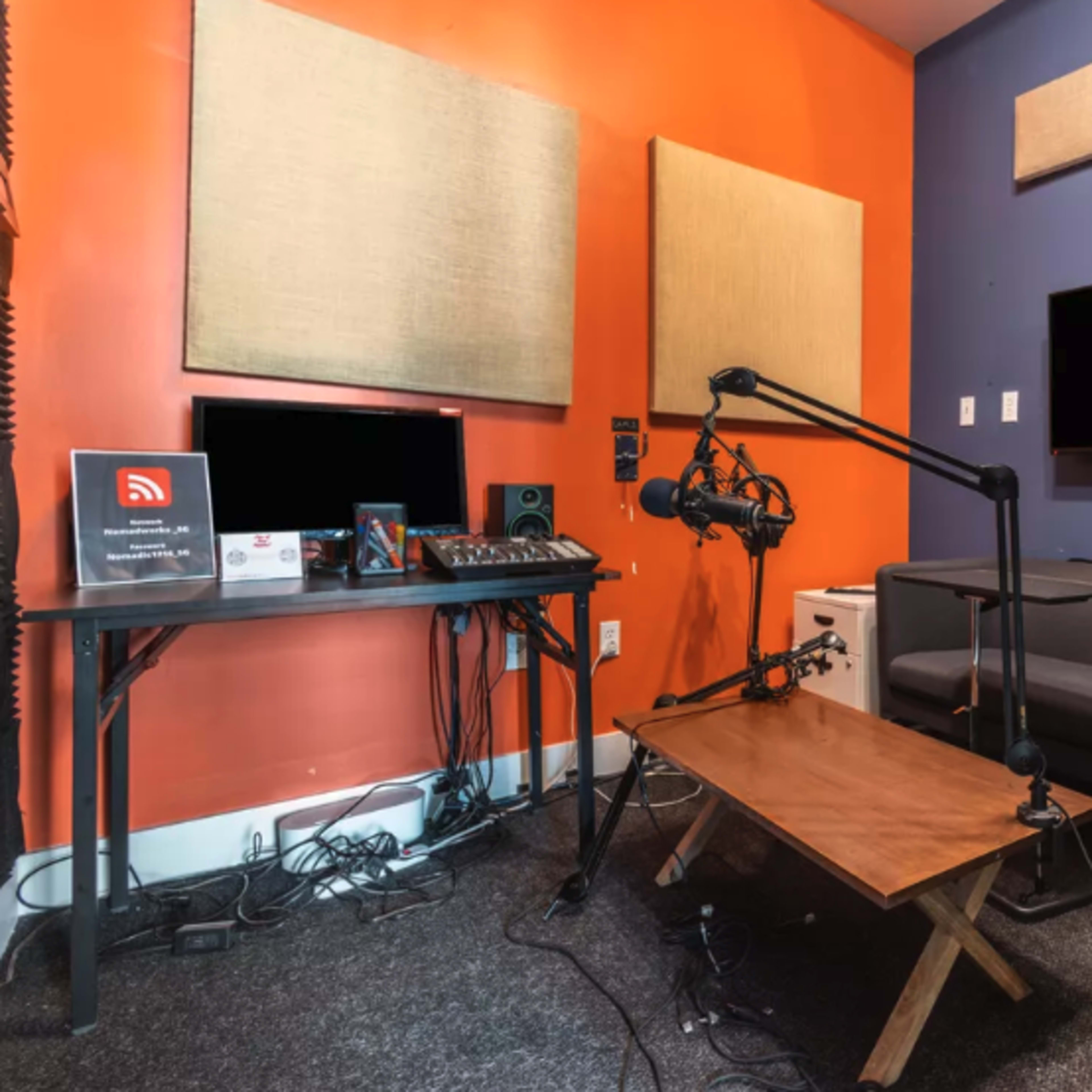 A small recording studio with an orange and gray color scheme, featuring a couch, microphone setup, a computer workstation, and acoustic panels on the walls.