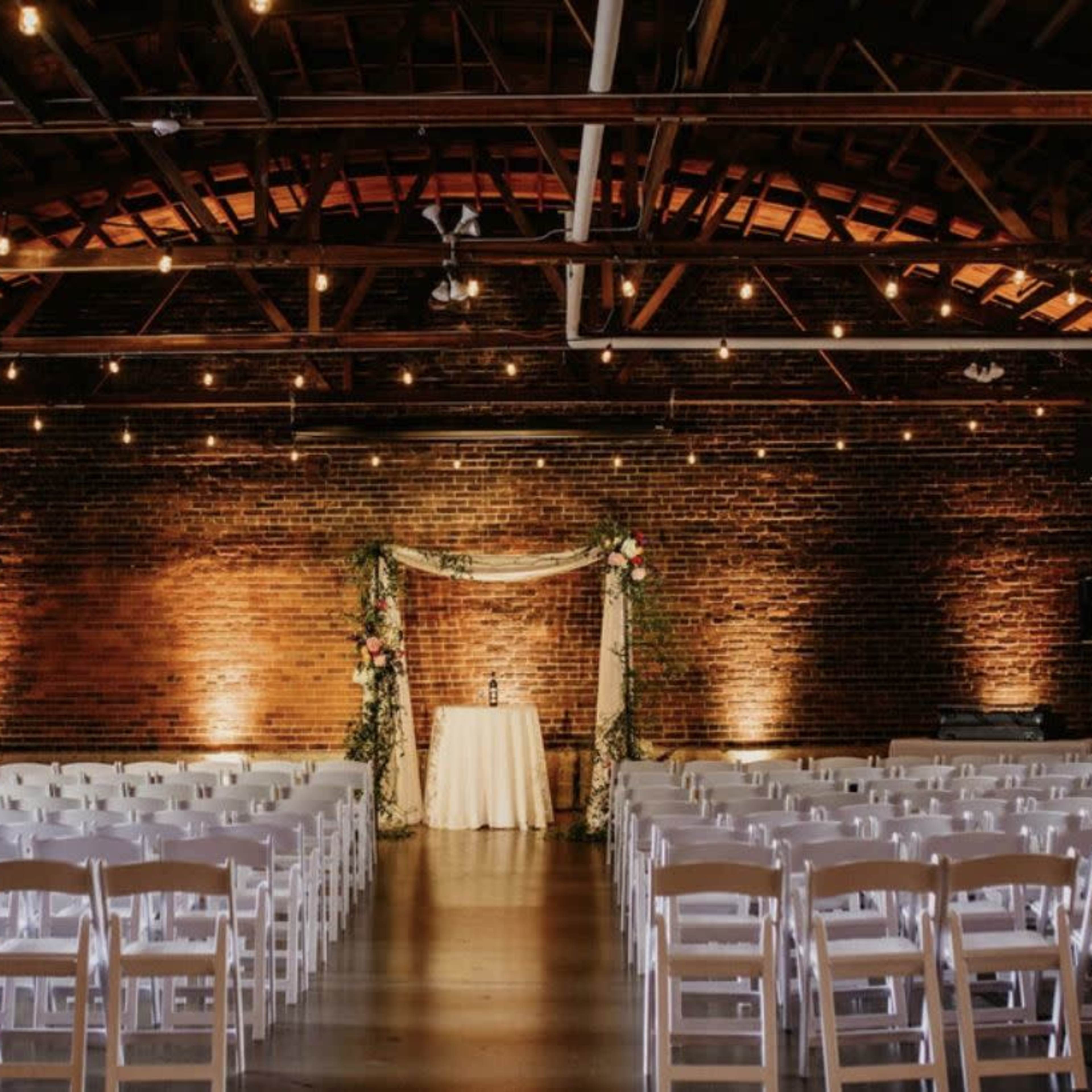 A wedding ceremony setup features rows of white chairs facing a decorated altar against a brick wall in a softly lit venue.