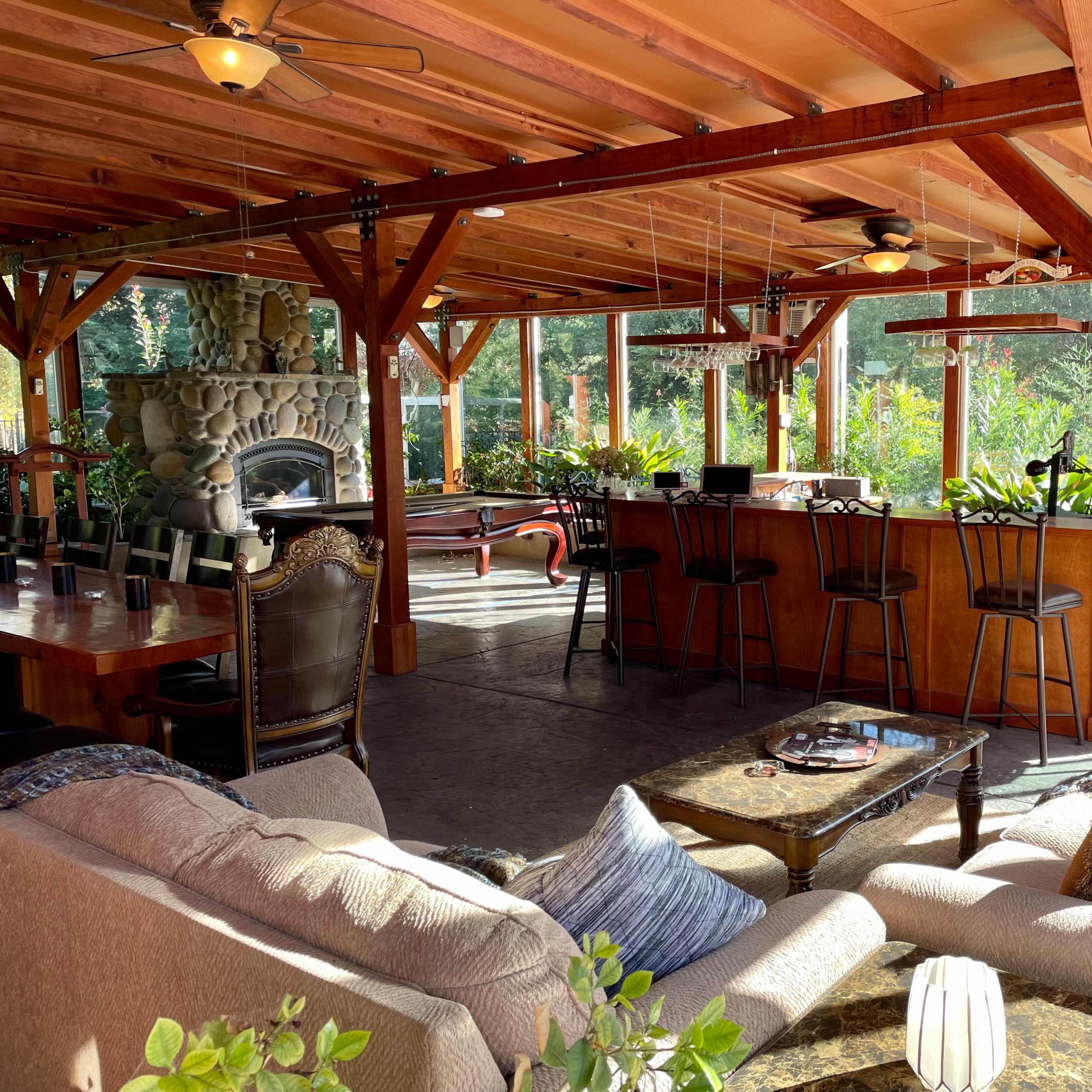 A spacious open-air room featuring a stone fireplace, a bar with stools, and seating areas with large windows overlooking a garden.