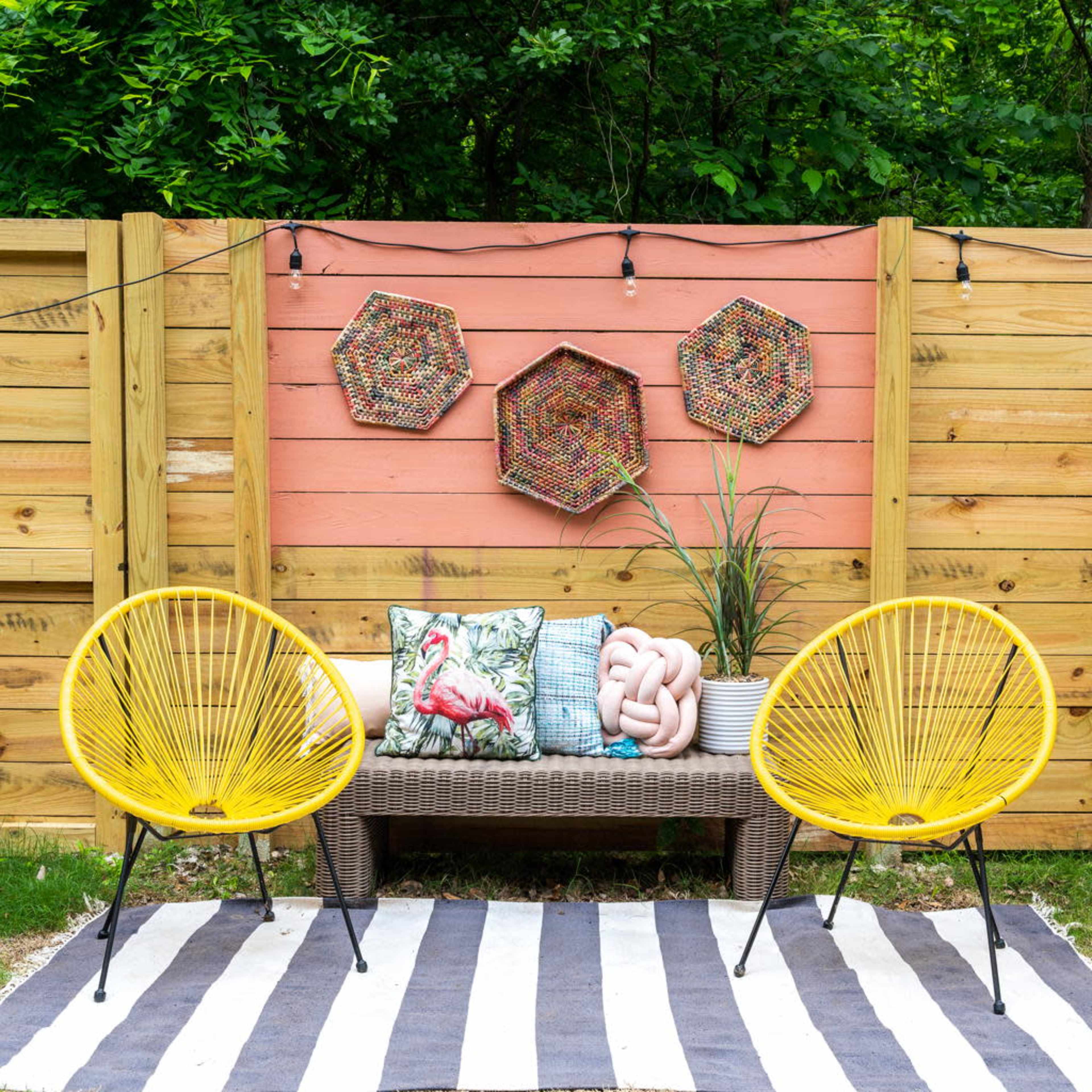 A small outdoor seating area with two bright yellow chairs, a bench with decorative pillows, and wall art against a wooden fence, all set on a striped rug in a green garden.
