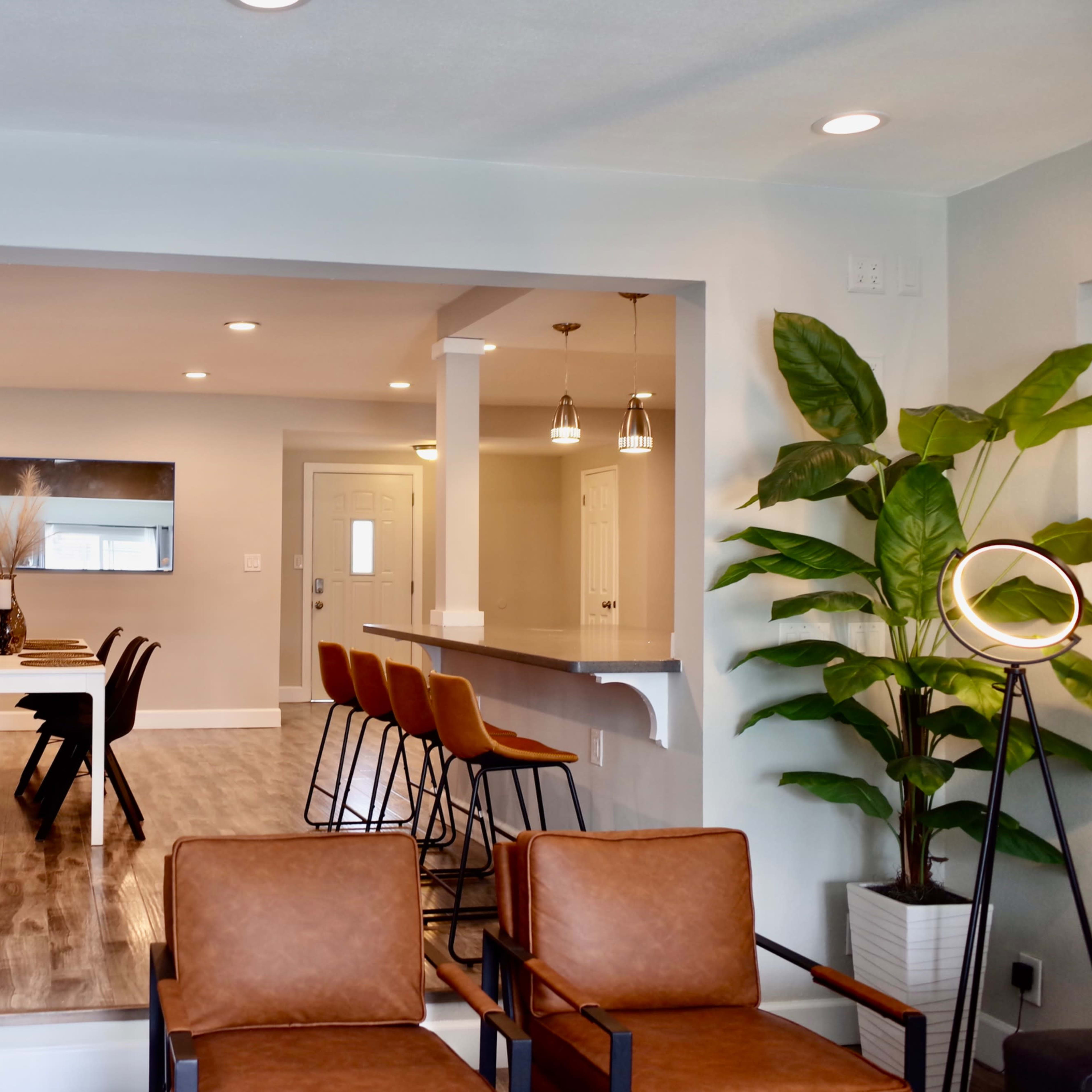A modern living space features a dining area with a wooden table and chairs, a kitchen bar with stools, and a lounge area with two brown chairs.