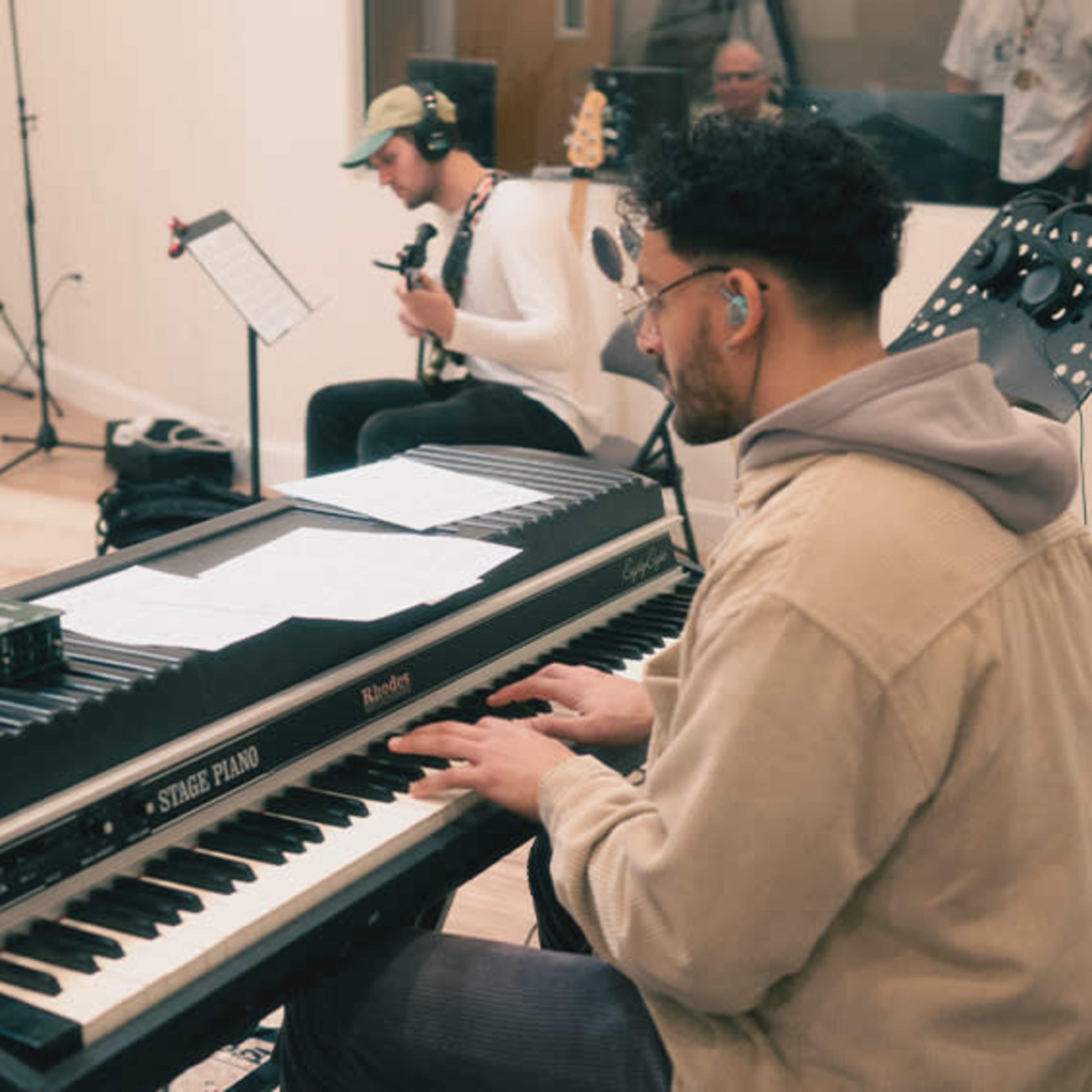 A musician plays a keyboard in a recording studio while another musician sits in the background with a guitar.