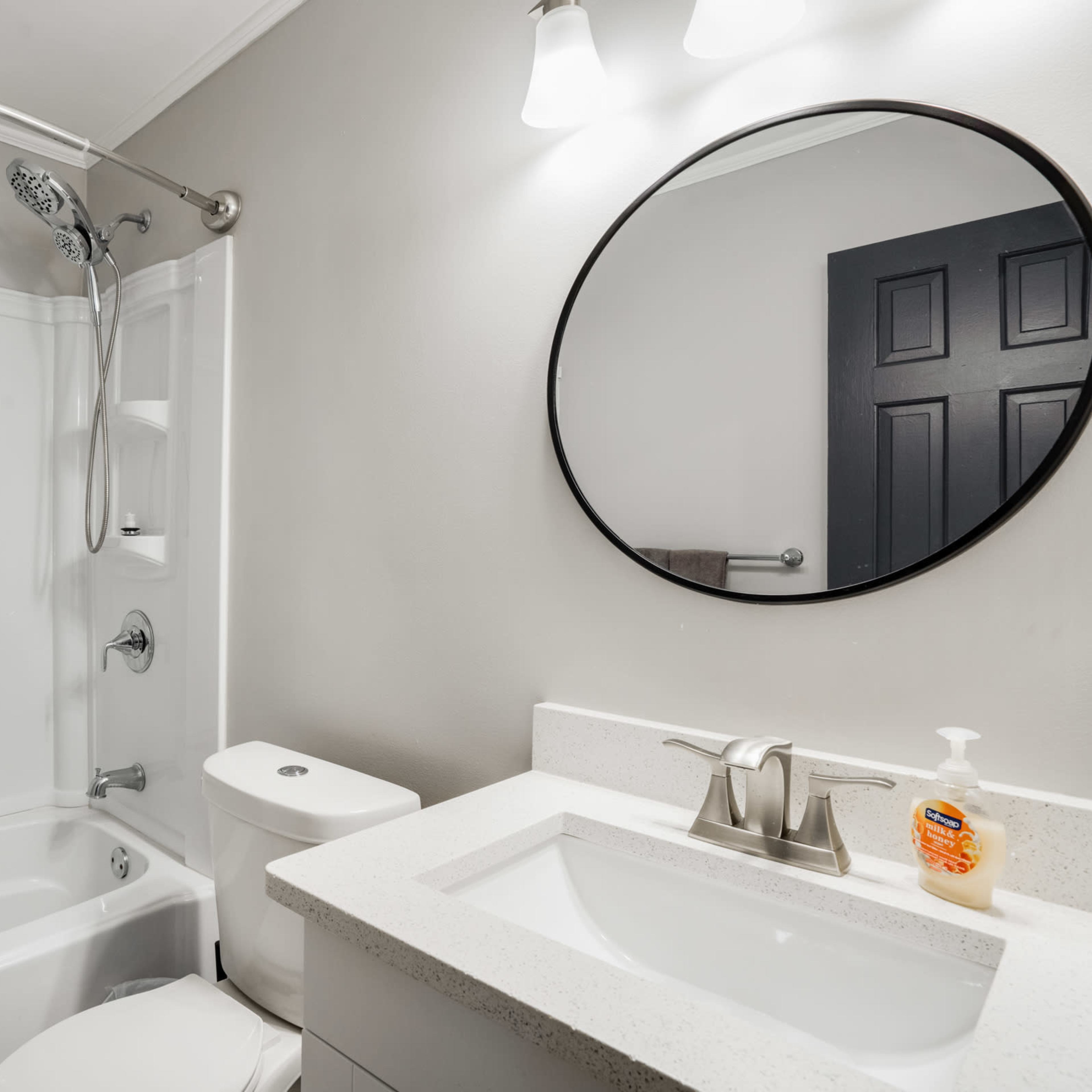 A modern bathroom featuring a shower-tub combo, a toilet, and a sink with a round mirror above it.