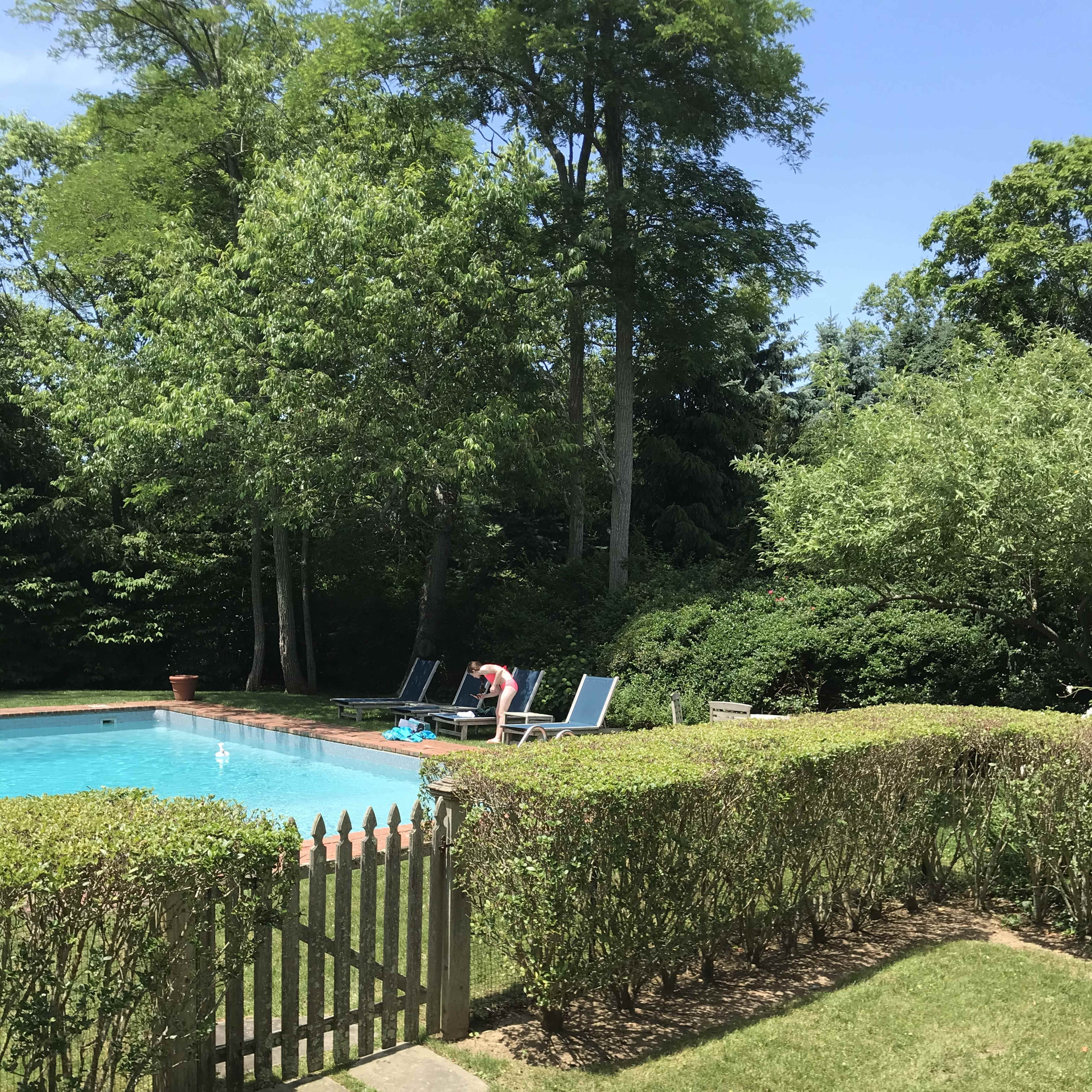 A person is lounging on a chair by a swimming pool surrounded by trees and hedges.