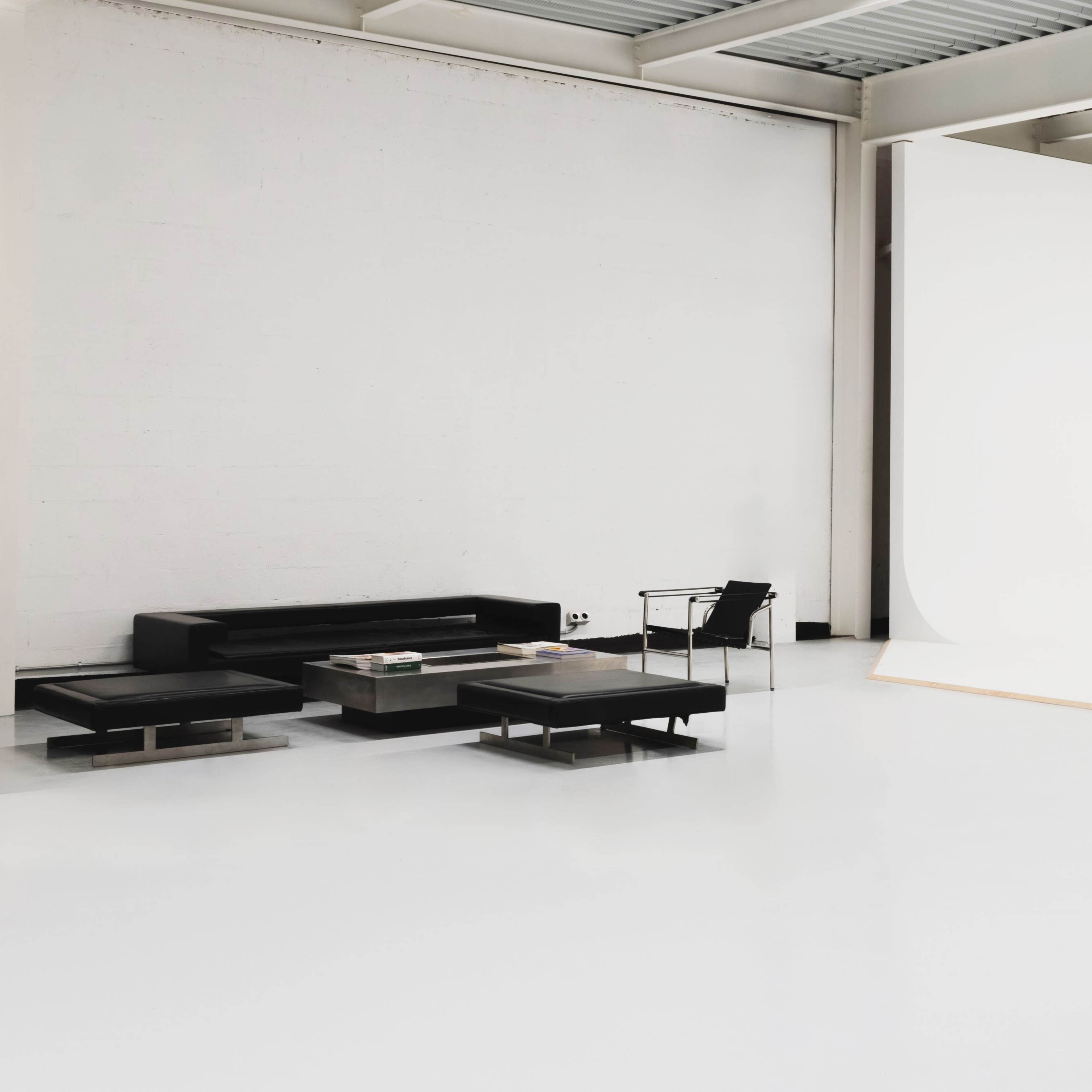 A modern, minimalist studio features a black sofa, a glass coffee table, and a large white backdrop.