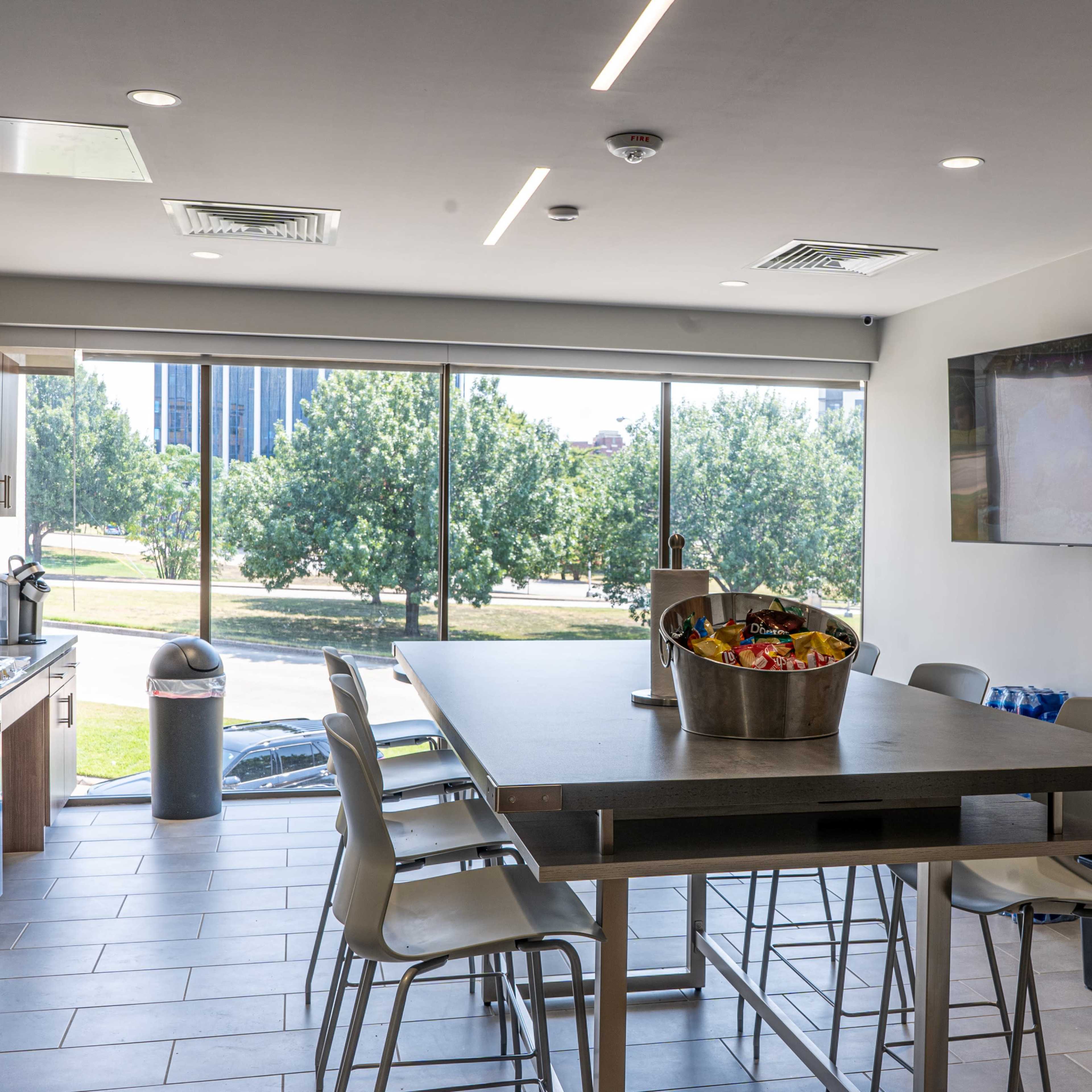 A modern kitchen features a large table with metal chairs, stainless steel appliances, and large windows showcasing a view of trees outside.