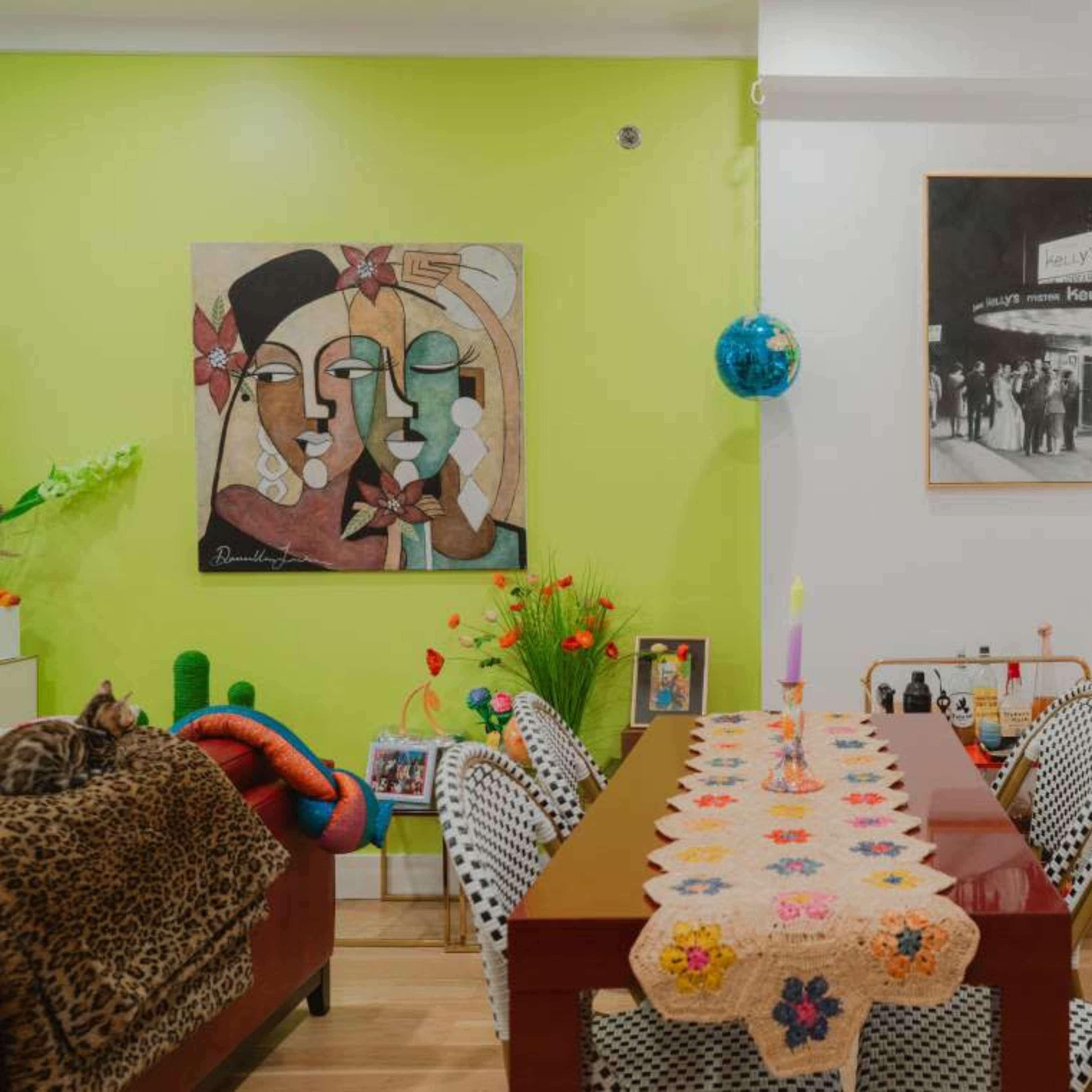 A colorful dining area with a table covered in a crocheted tablecloth, surrounded by decorative chairs, and a wall adorned with artwork and shelves displaying various items.