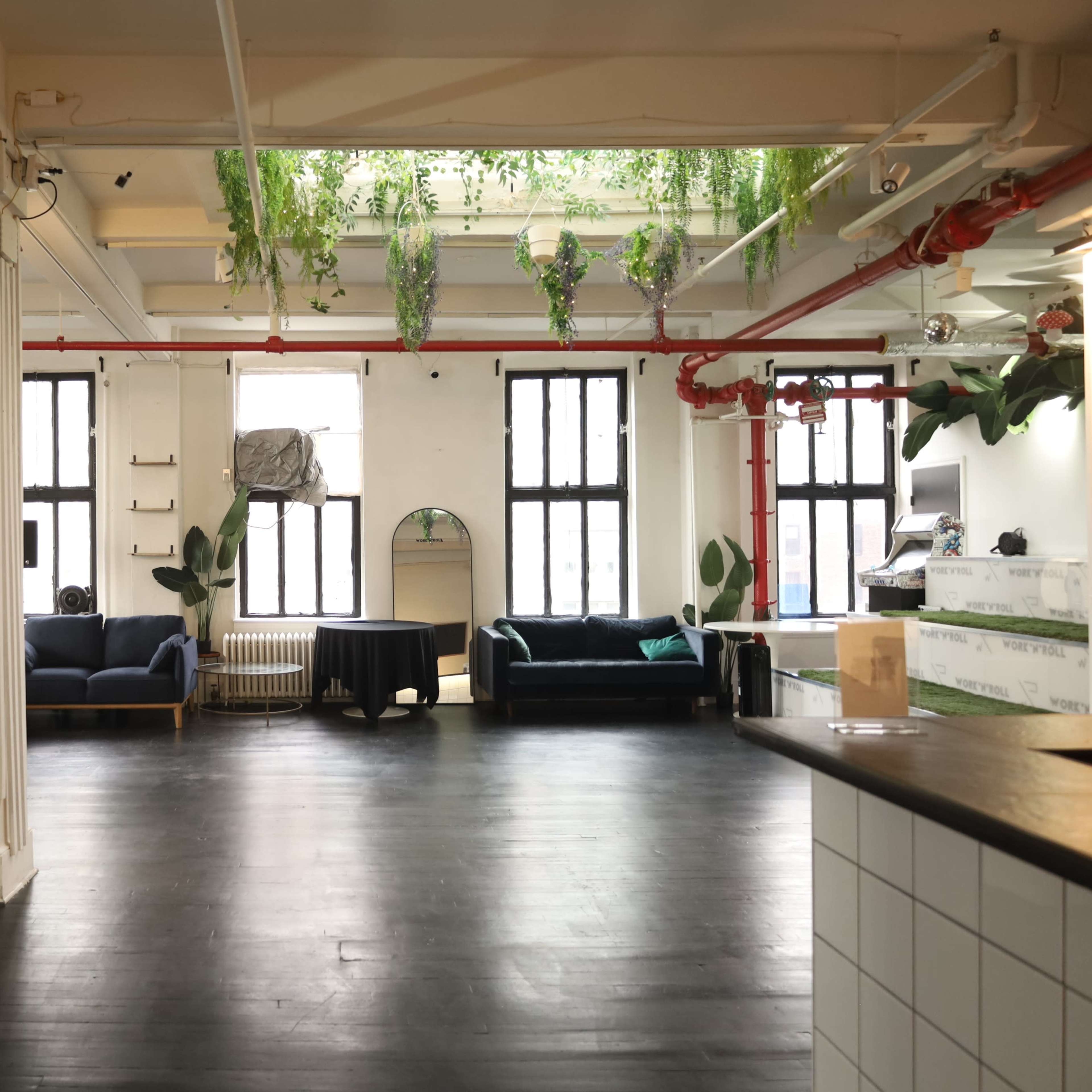 An open, airy space with large windows, featuring green plants hanging from the ceiling and a mix of seating arrangements, including two sofas and a small table.