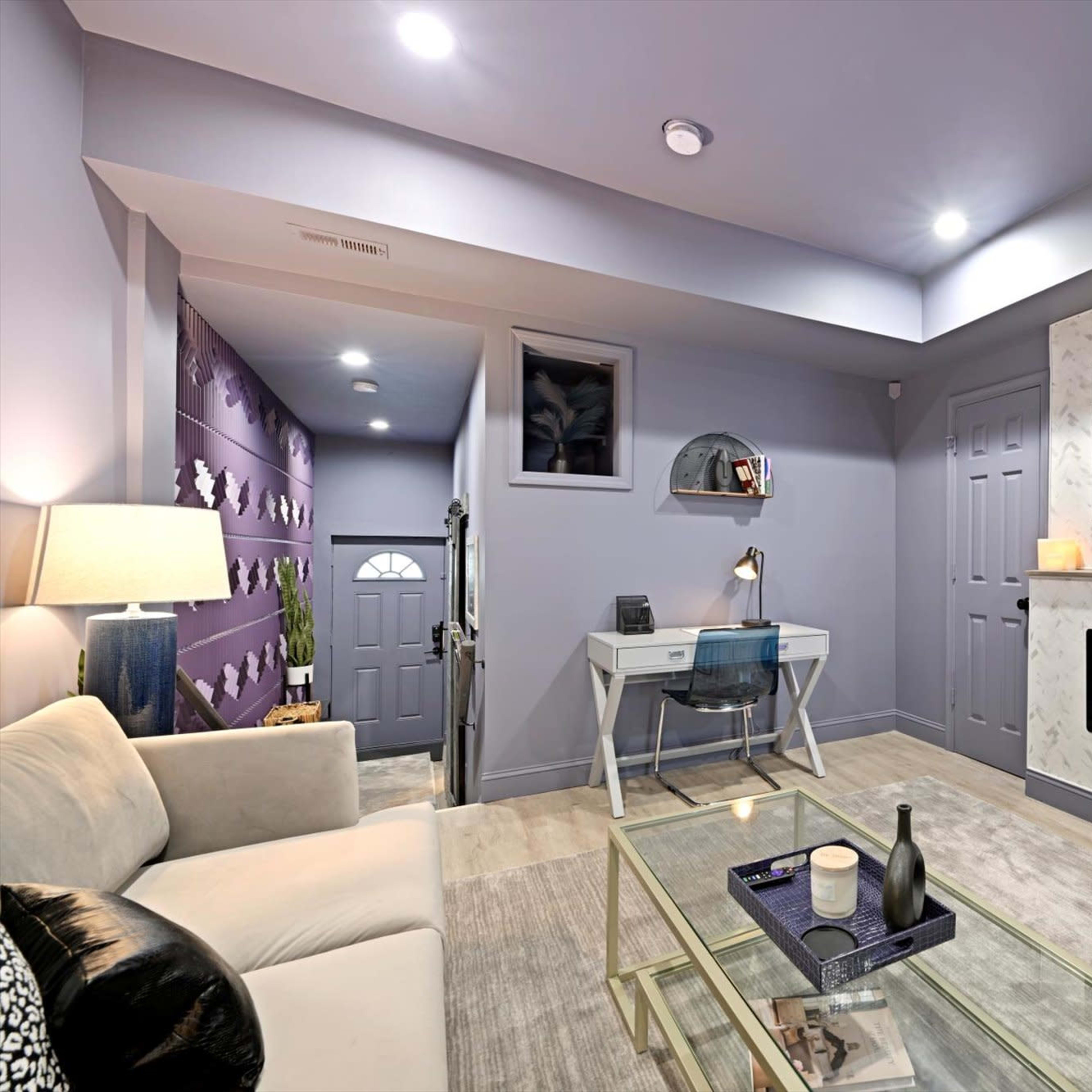 A modern living room with a white sofa, a glass coffee table, an electric fireplace beneath a television, and a desk against a purple accent wall.