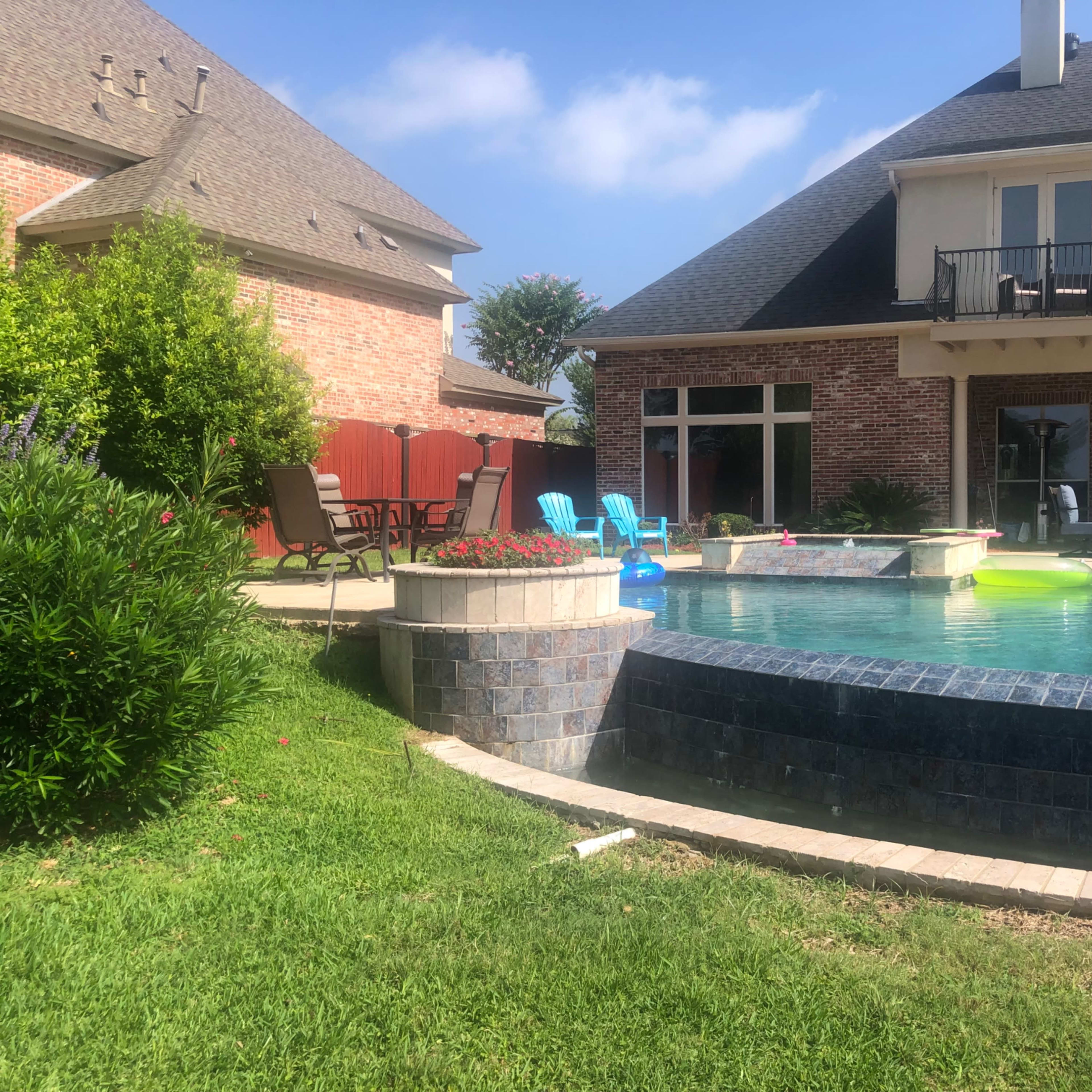 A backyard pool area with a brick house, lounge chairs, and colorful inflatable floaties in the water.