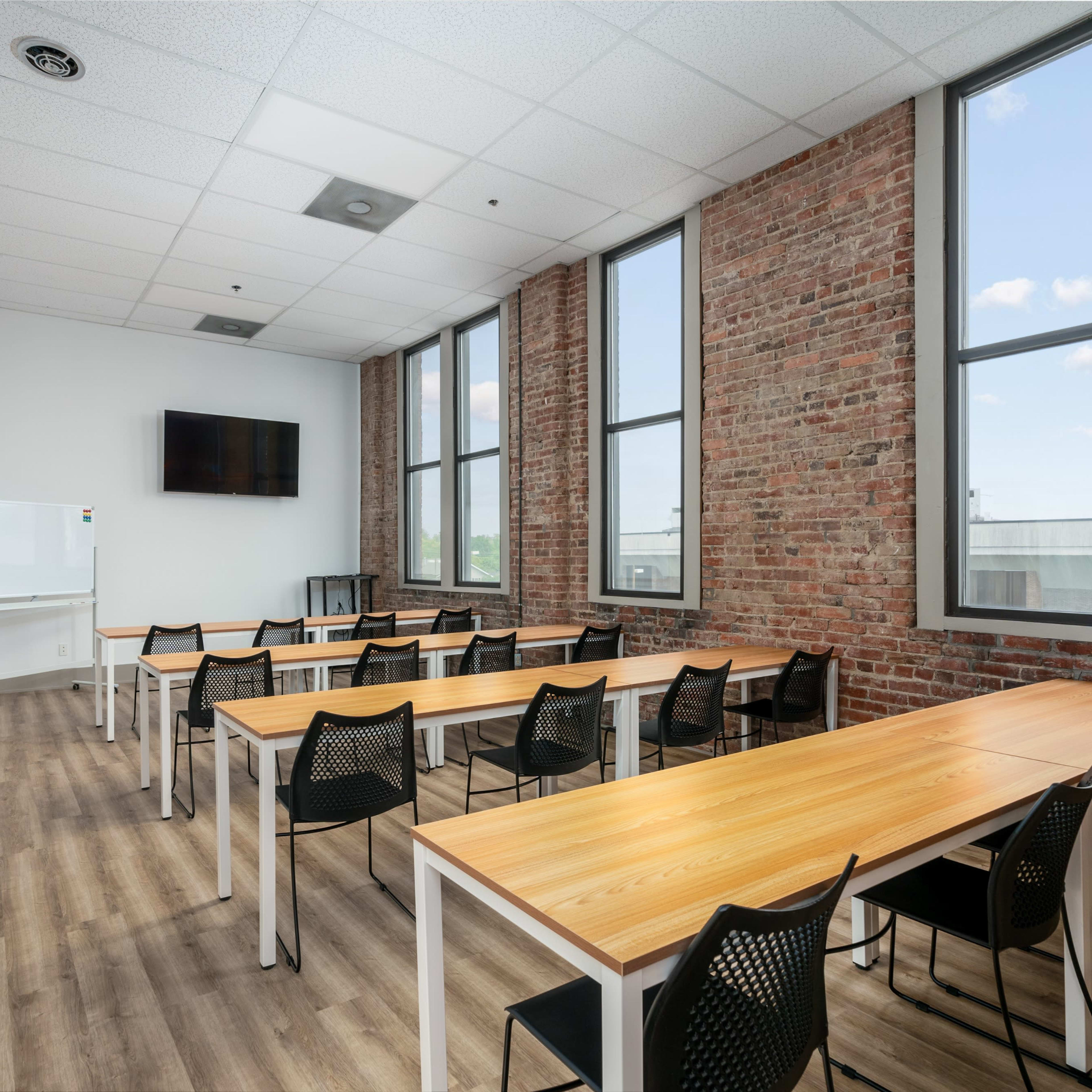 A modern classroom with wooden tables and black chairs, large windows, and exposed brick walls.