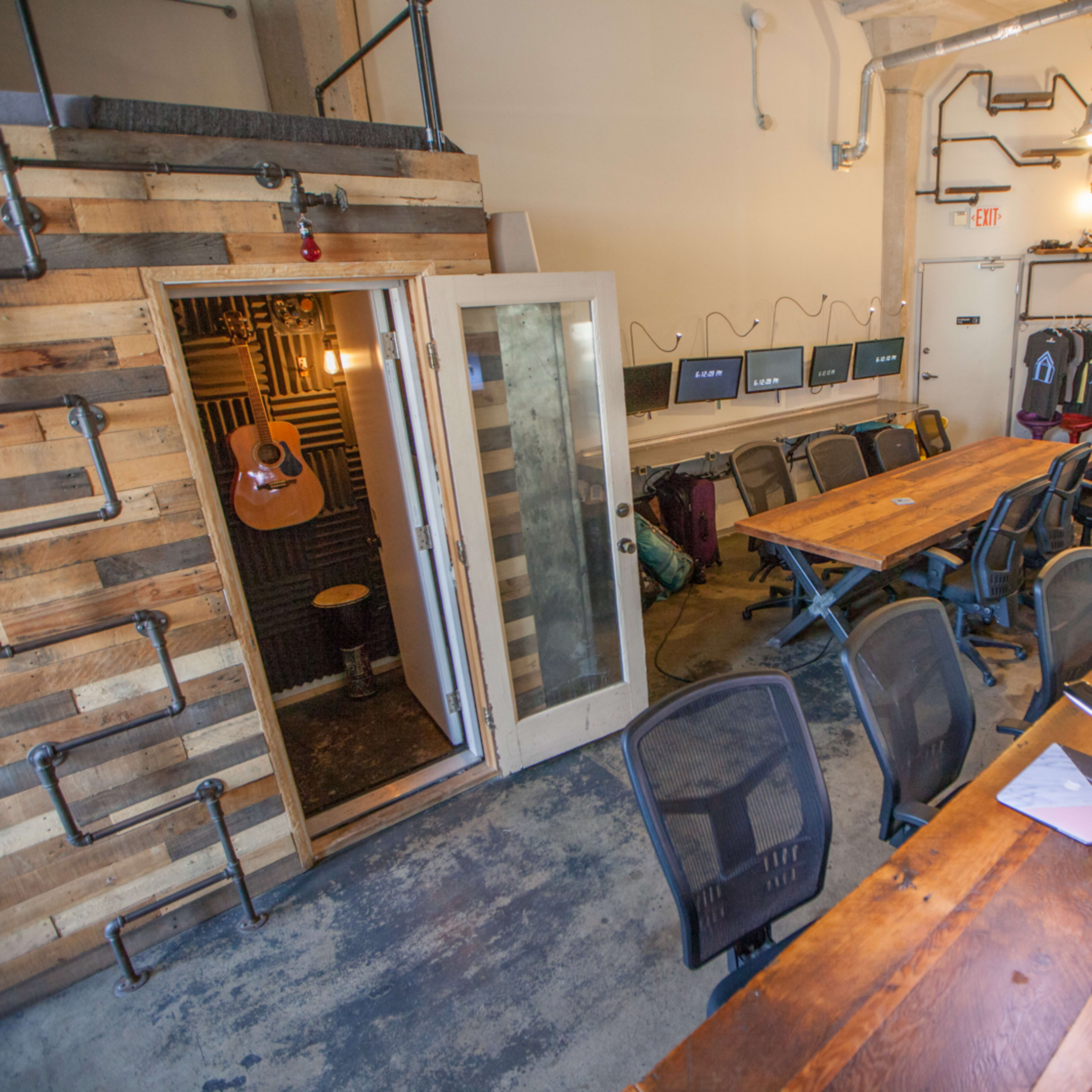A modern workspace with a wooden conference table, office chairs, and a small soundproof room with an acoustic guitar inside.