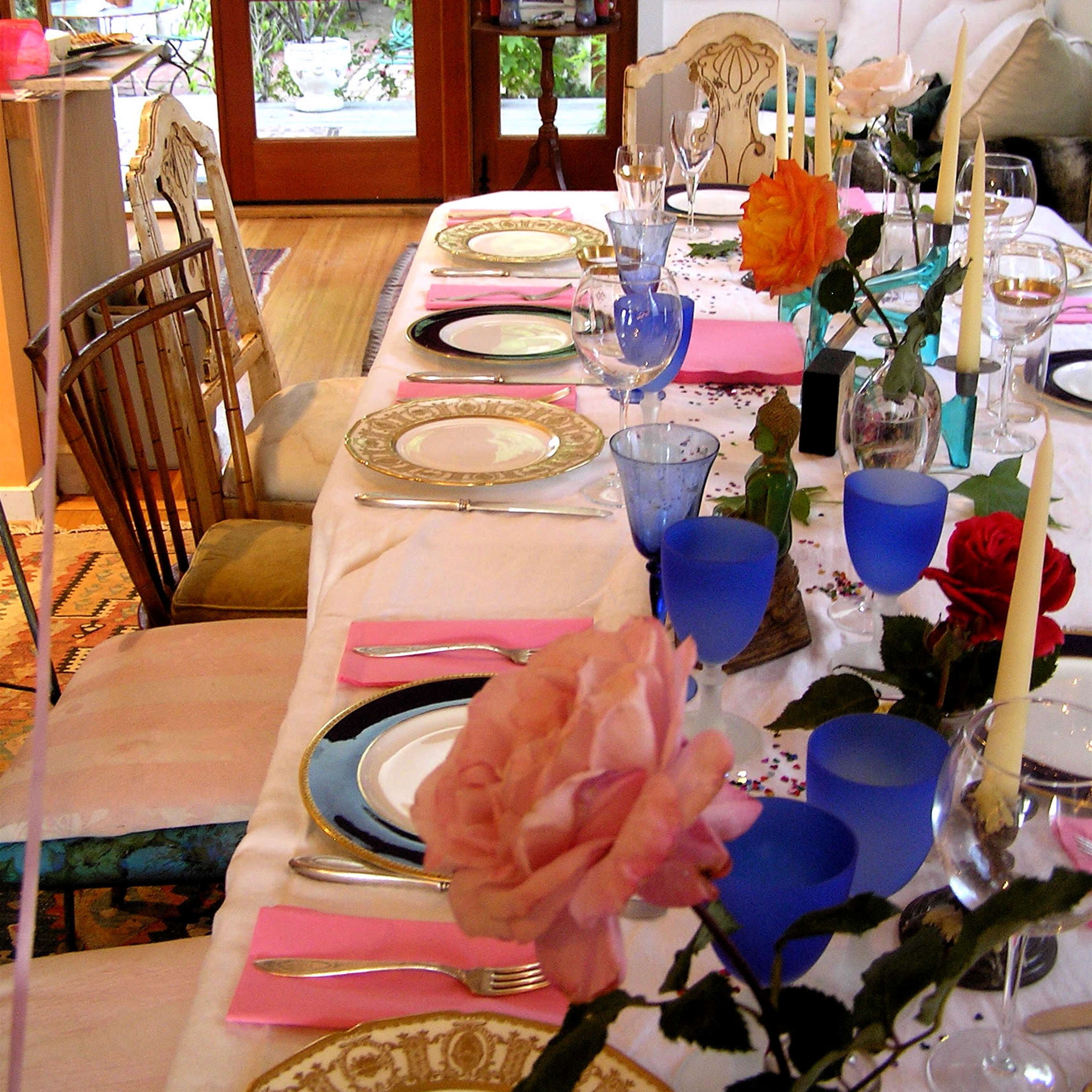 A dining table is set with plates, glasses, and floral decorations, arranged in a well-lit room overlooking a garden through large windows.