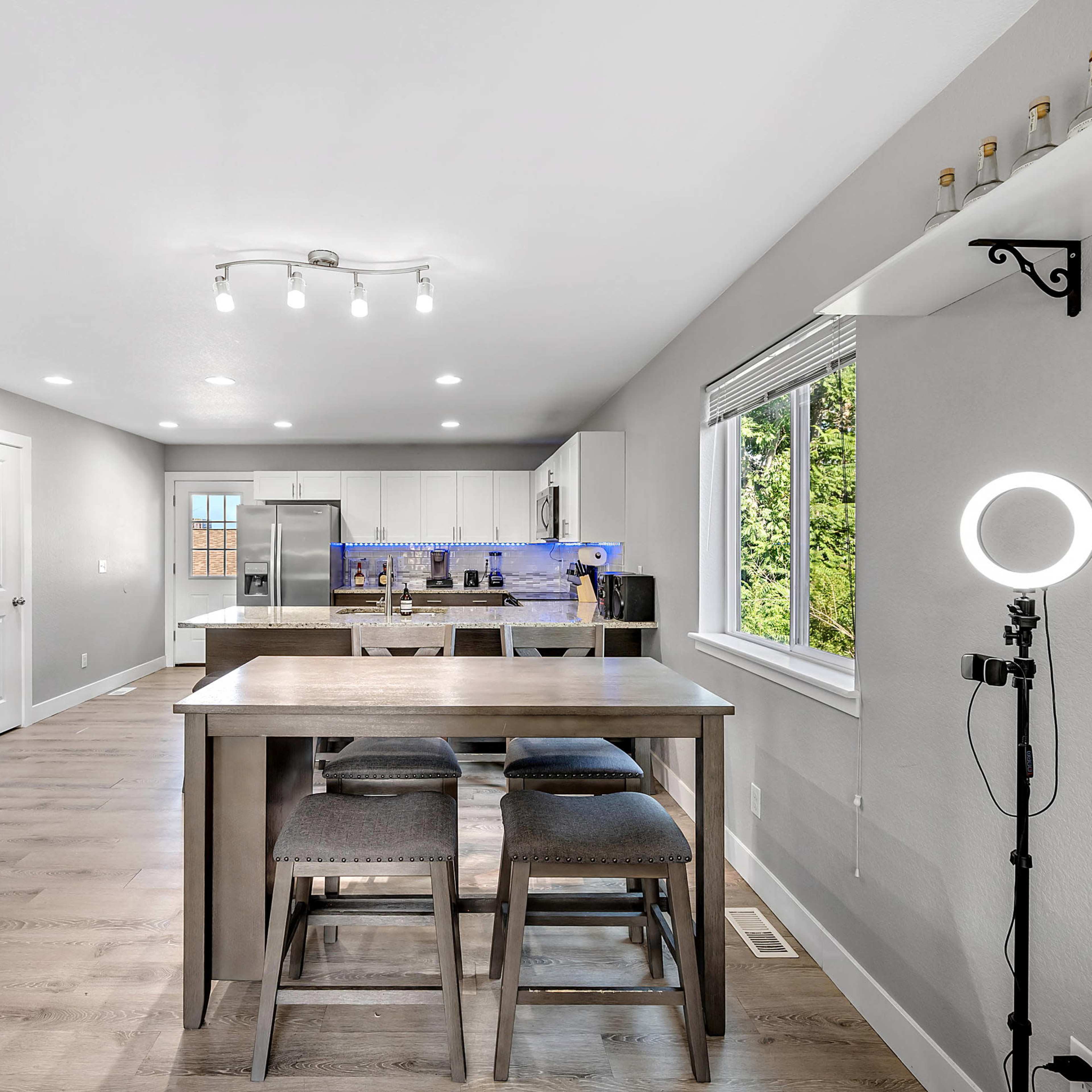A modern kitchen with a dining table and four wooden chairs, featuring stainless steel appliances and large windows.