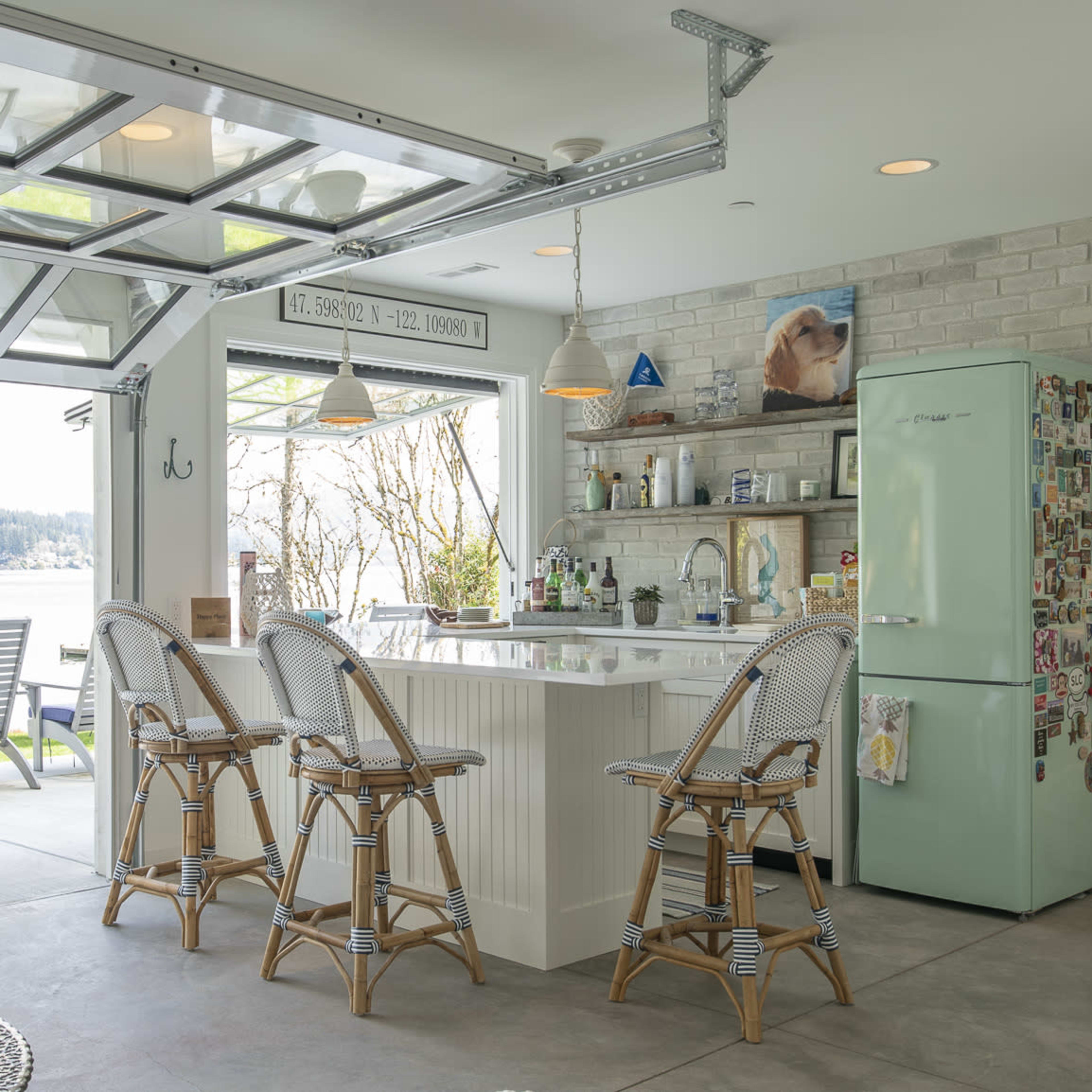 A modern kitchen with a bar and four rattan stools opens to a scenic view of a body of water through large glass doors.